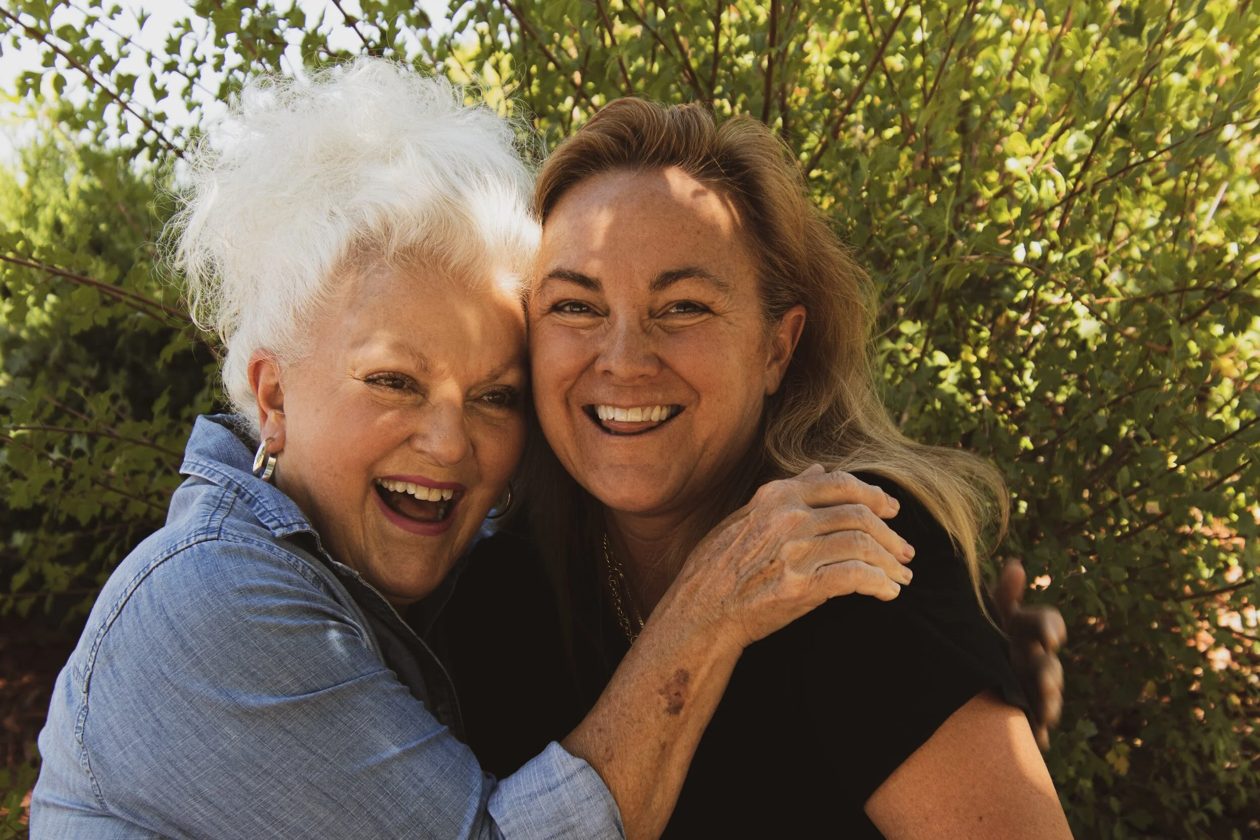 Two smiling women hugging outdoors in front of green foliage.