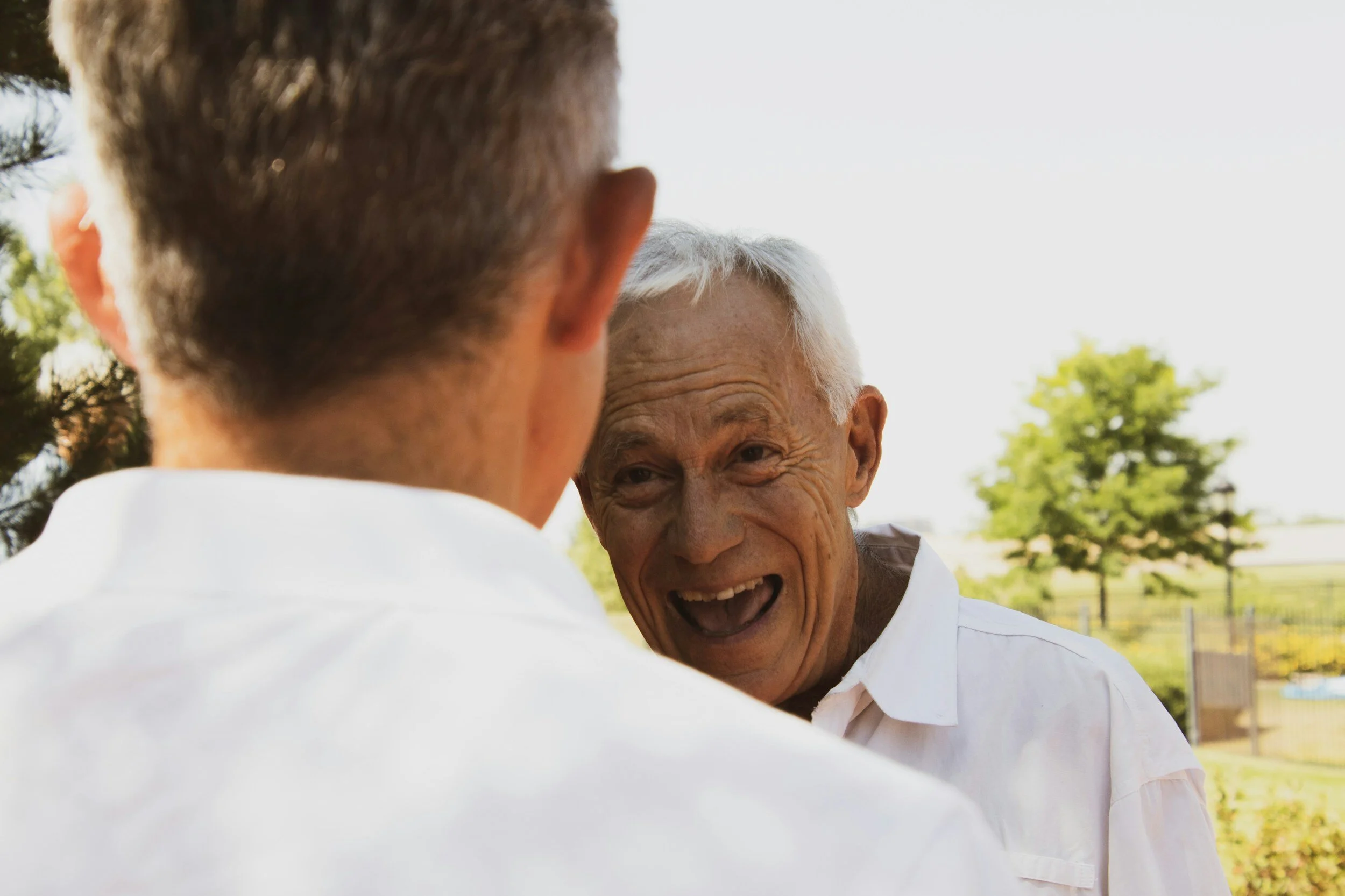 Two elderly men smiling and conversing outside on a sunny day, one facing the camera, the other with his back turned.