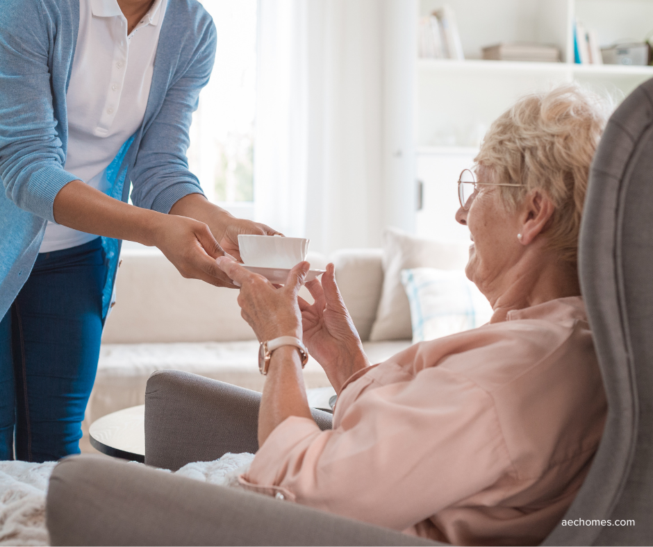 Caregiver handing a cup to an elderly woman seated in a living room.