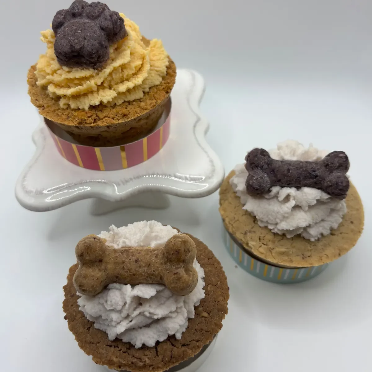 Three cupcakes decorated with frosting and dog biscuit-shaped treats, displayed on a white surface and a small white stand.