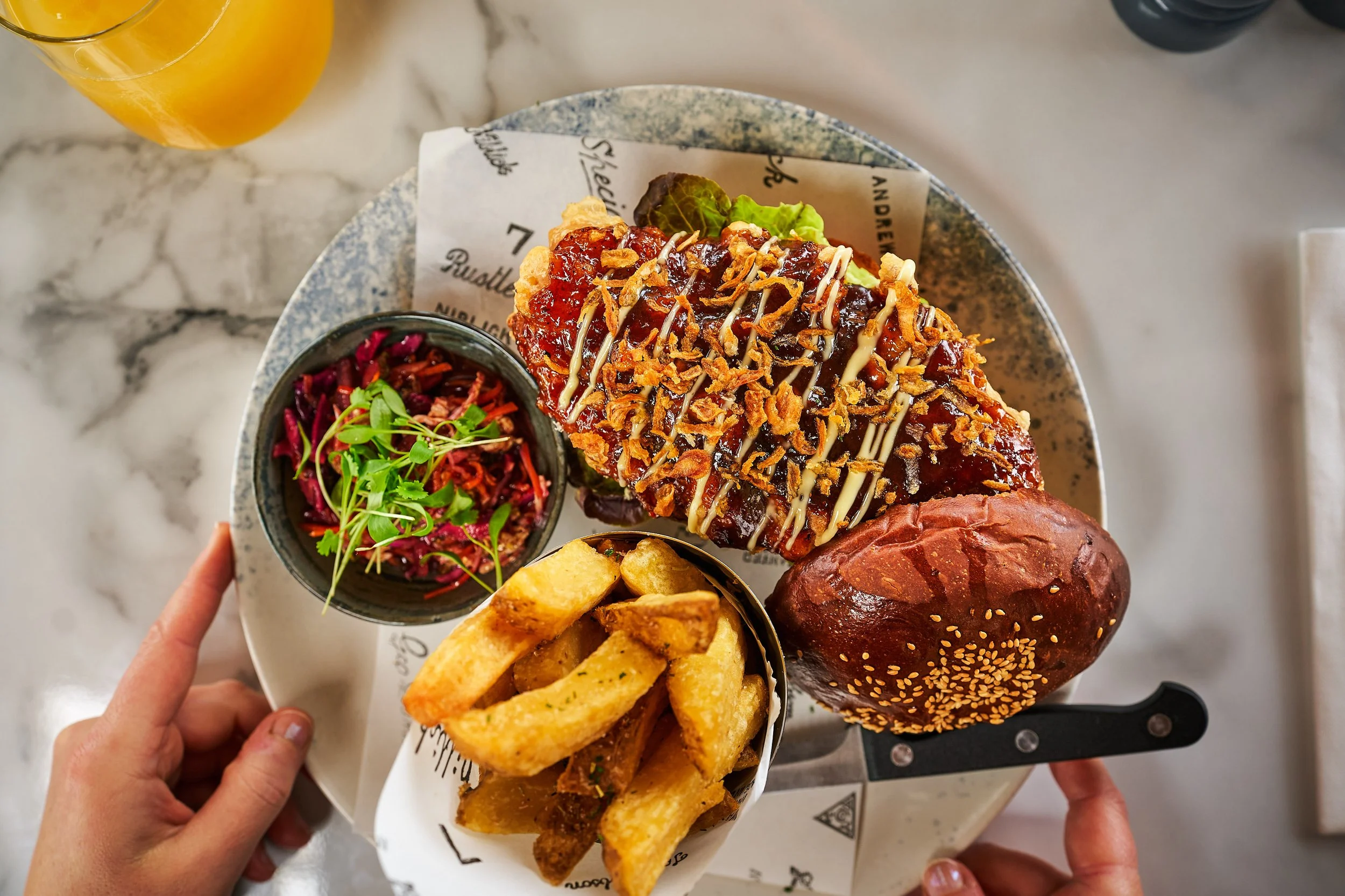 An open bun with a chicken burger topped with sauce, a side of chips, a small bowl of ginger slaw with greens, and a bread roll, all on a round plate on a white marble table, with a glass of orange juice nearby.