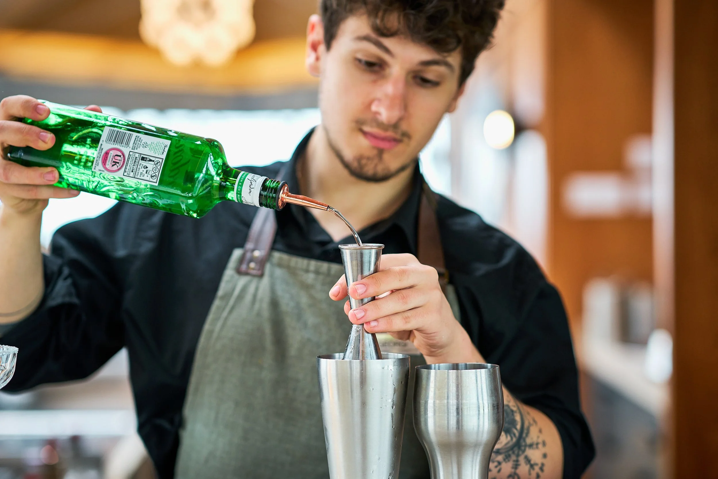 A bartender wearing a green apron pours alcohol into a cocktail shaker.