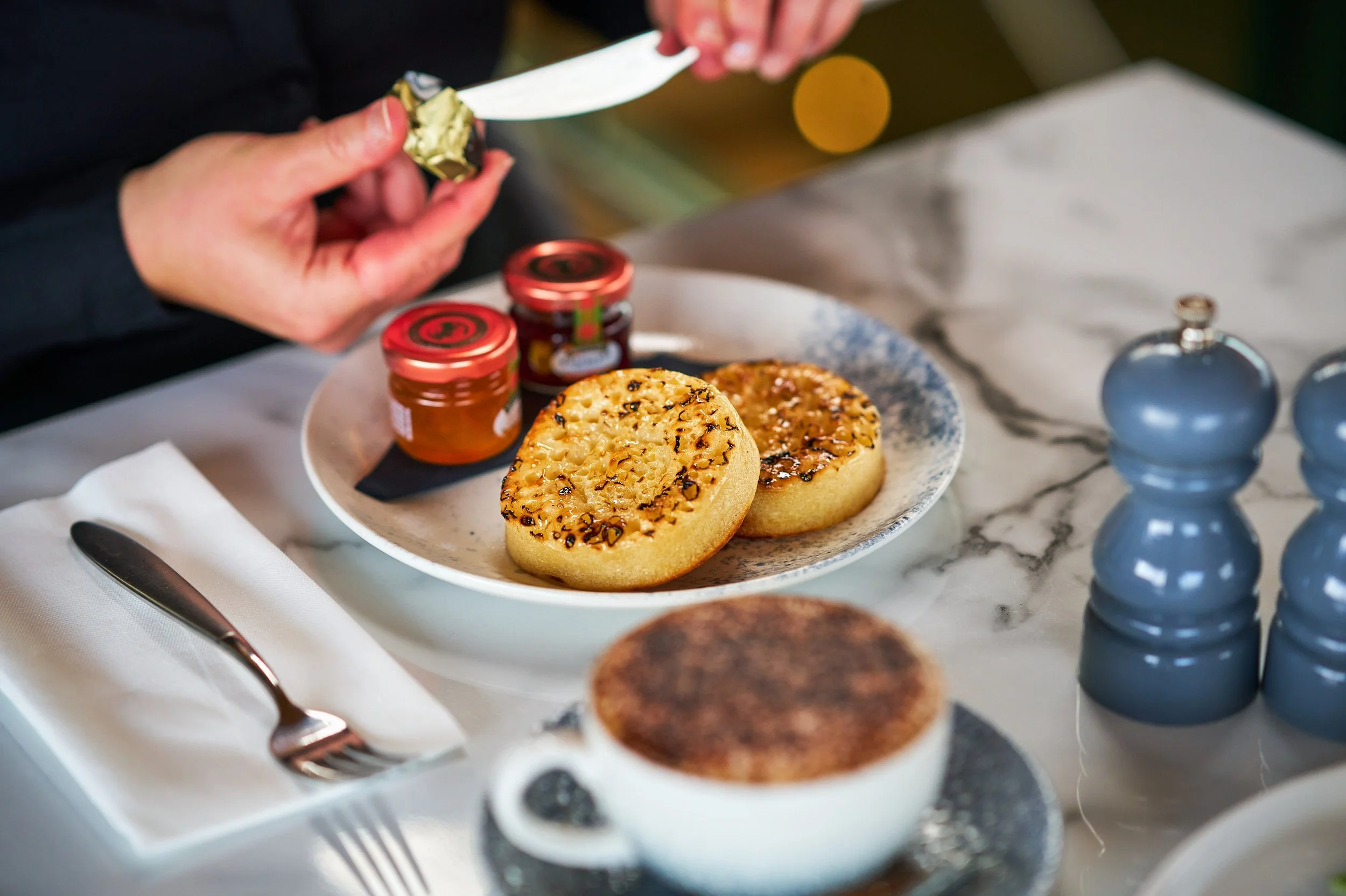 Person spreading butter on toasted scones served with jam on plates, on a marble table.