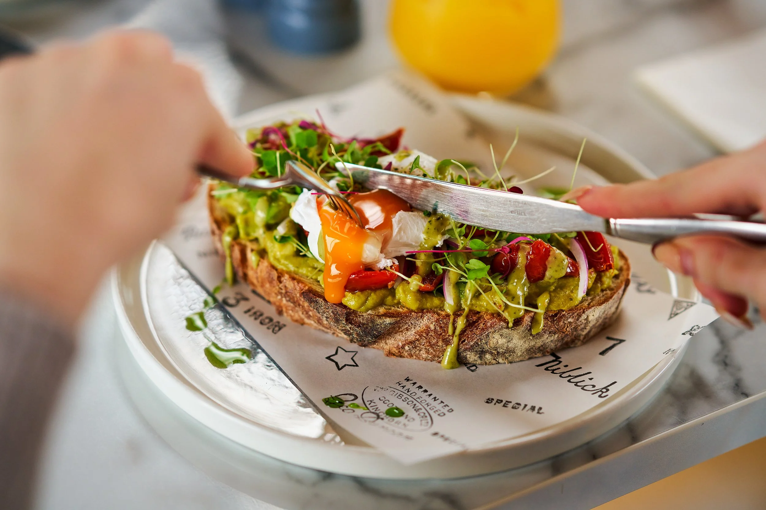 Person cutting into a colourful open-faced avocado toast topped with a poached egg and microgreens on a piece of toasted bread, served on a tray with a white paper liner.