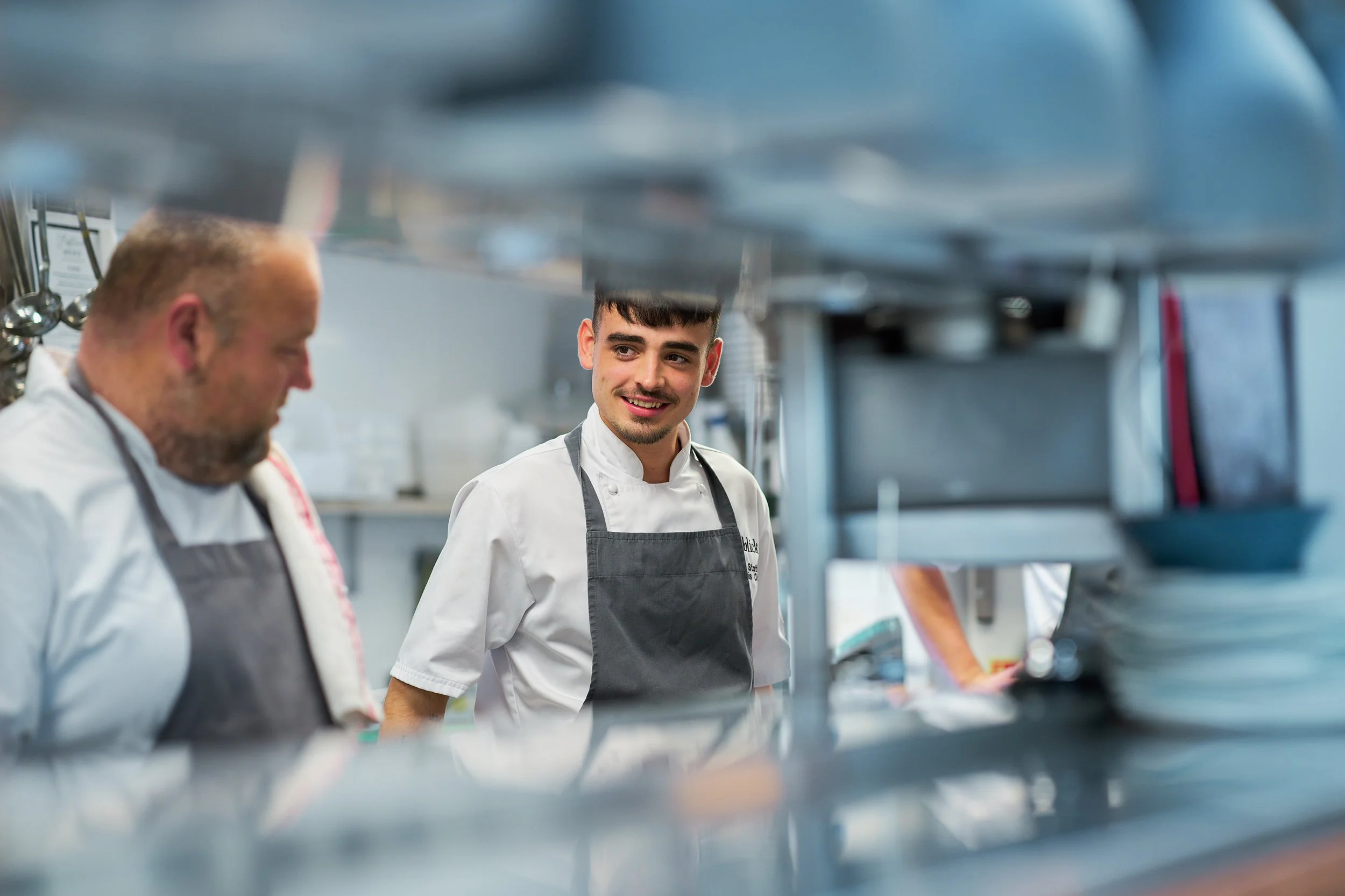 Two chefs working in a professional kitchen, one is a middle-aged man with a beard and the other is a young man with dark hair, both wearing white chef jackets and gray aprons.
