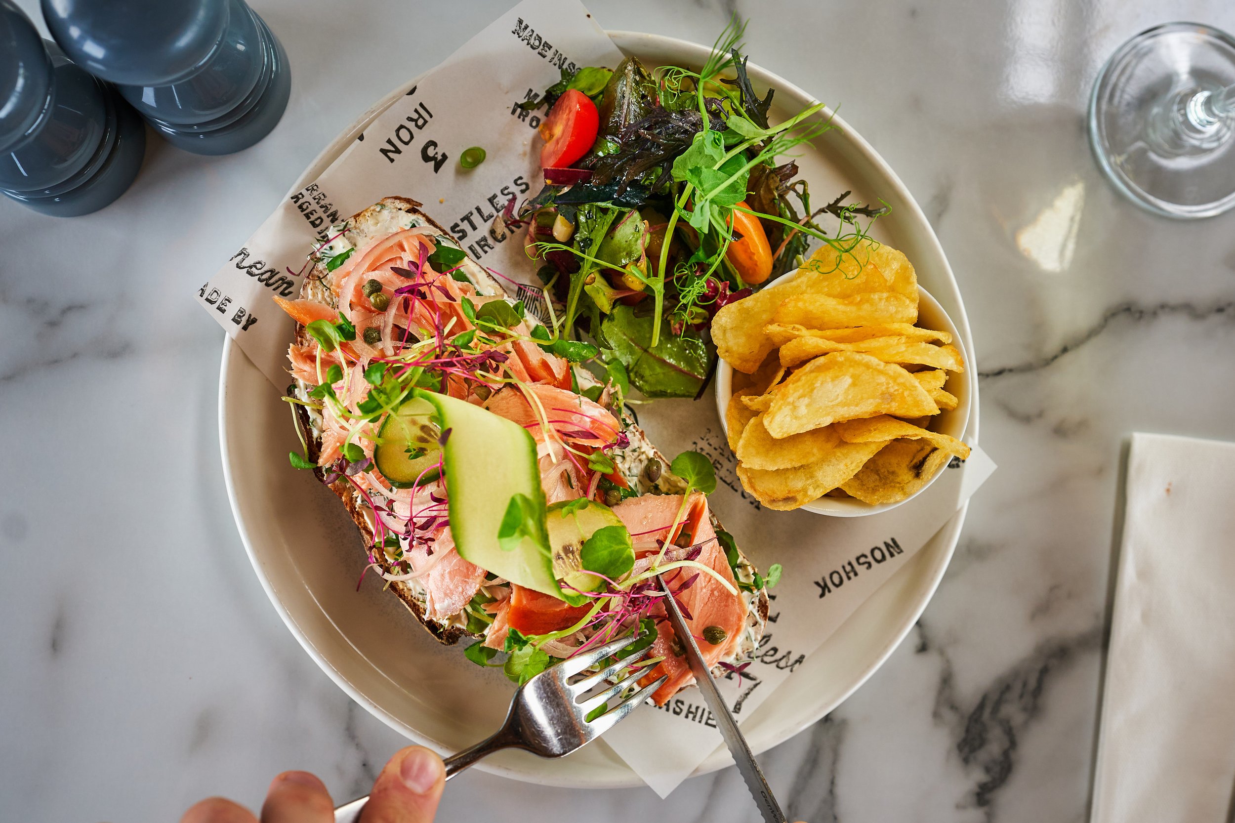 Plate with open-faced smoked salmon toast topped with microgreens, cucumber, and capers, served with a side salad and potato chips on a marble table.