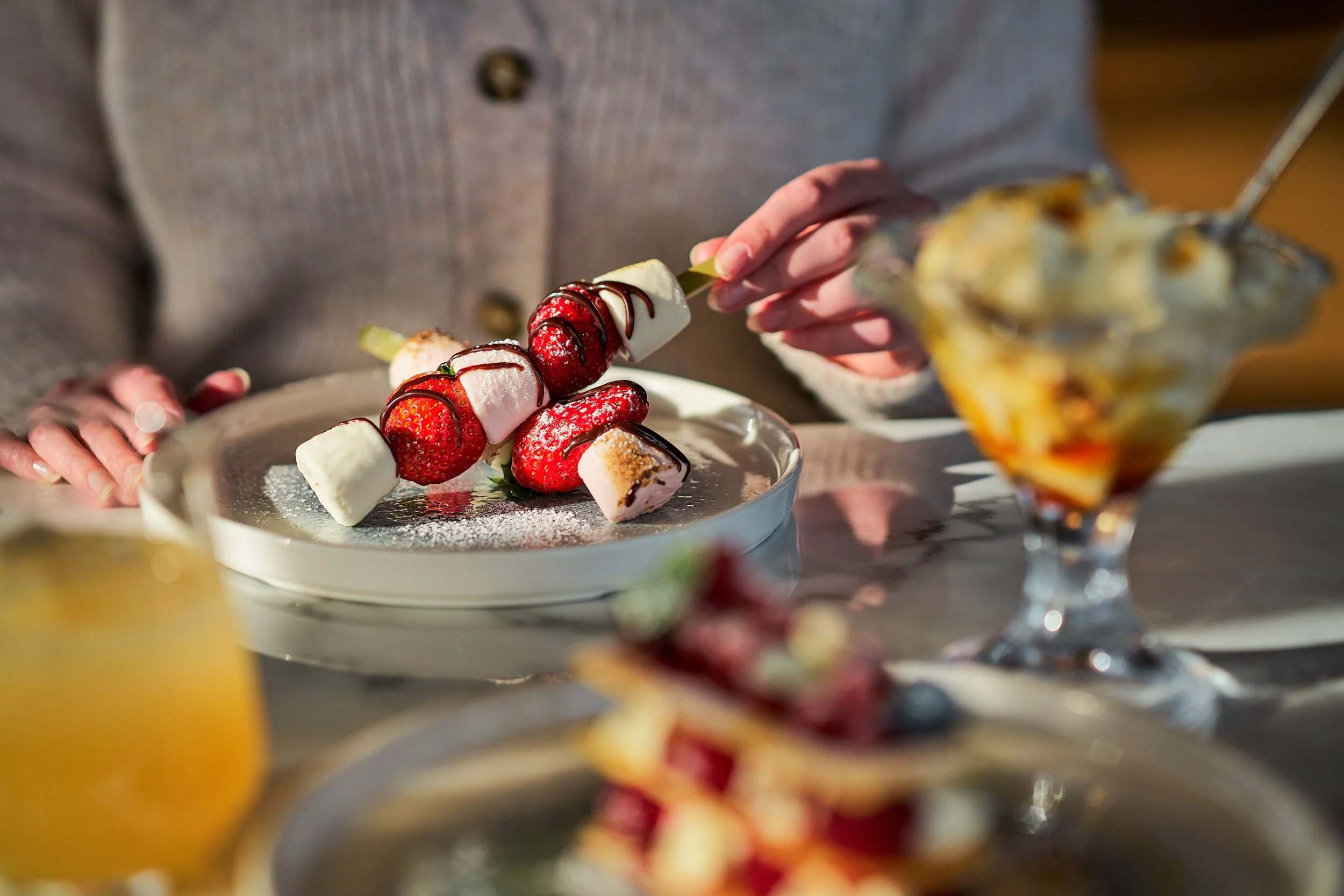 A hand holds a skewer with local Scottish strawberries and marshmallows drizzled in chocolate sauce and dusted with icing sugar.