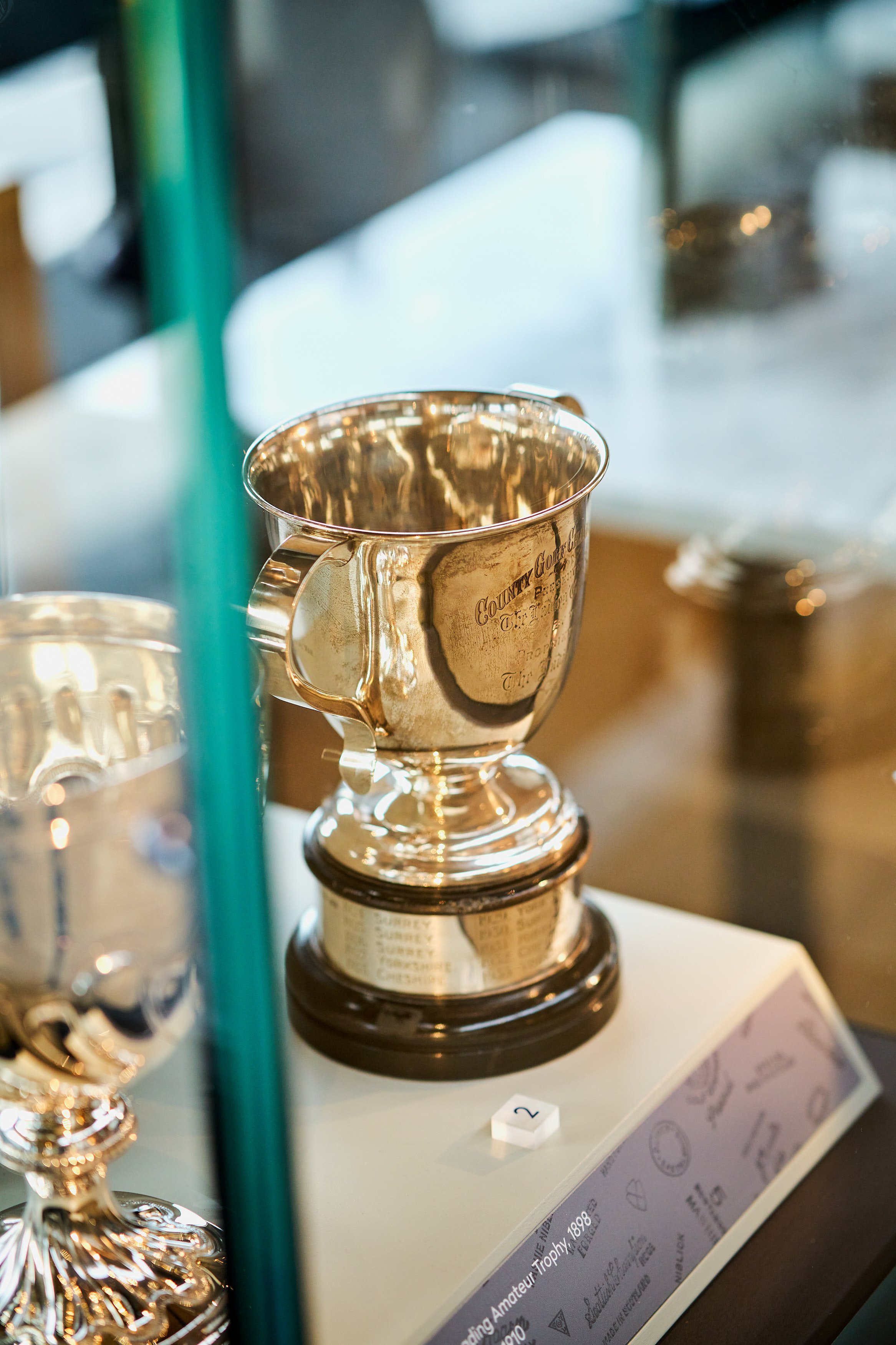 A silver trophy cup with engraved text, displayed on a white platform with a label, inside a glass case.