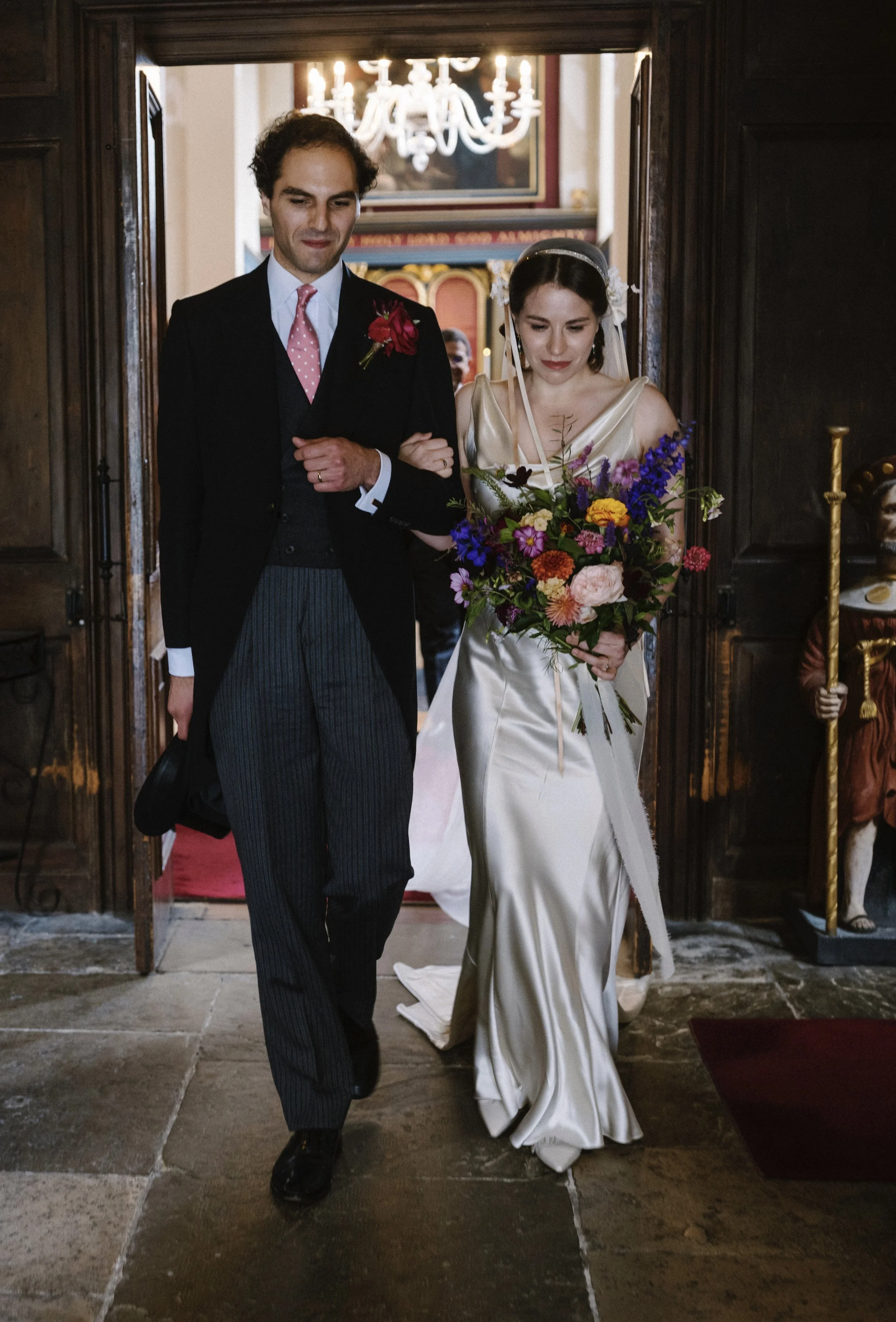 A bride in a cream satin gown holding a colorful bouquet walking with a groom dressed in a black tailcoat with a pink polka dot tie, inside a church with a chandelier and religious statues.
