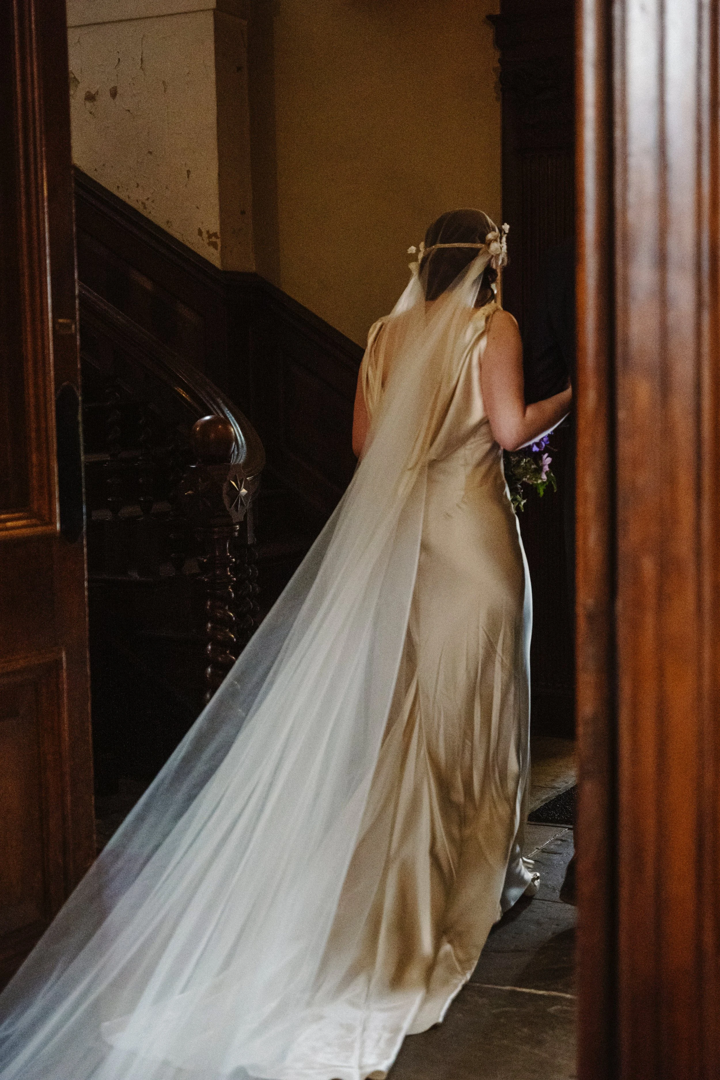 A bride in a satin wedding dress with a long Juliet Cap veil walking down a wooden staircase, viewed from behind.