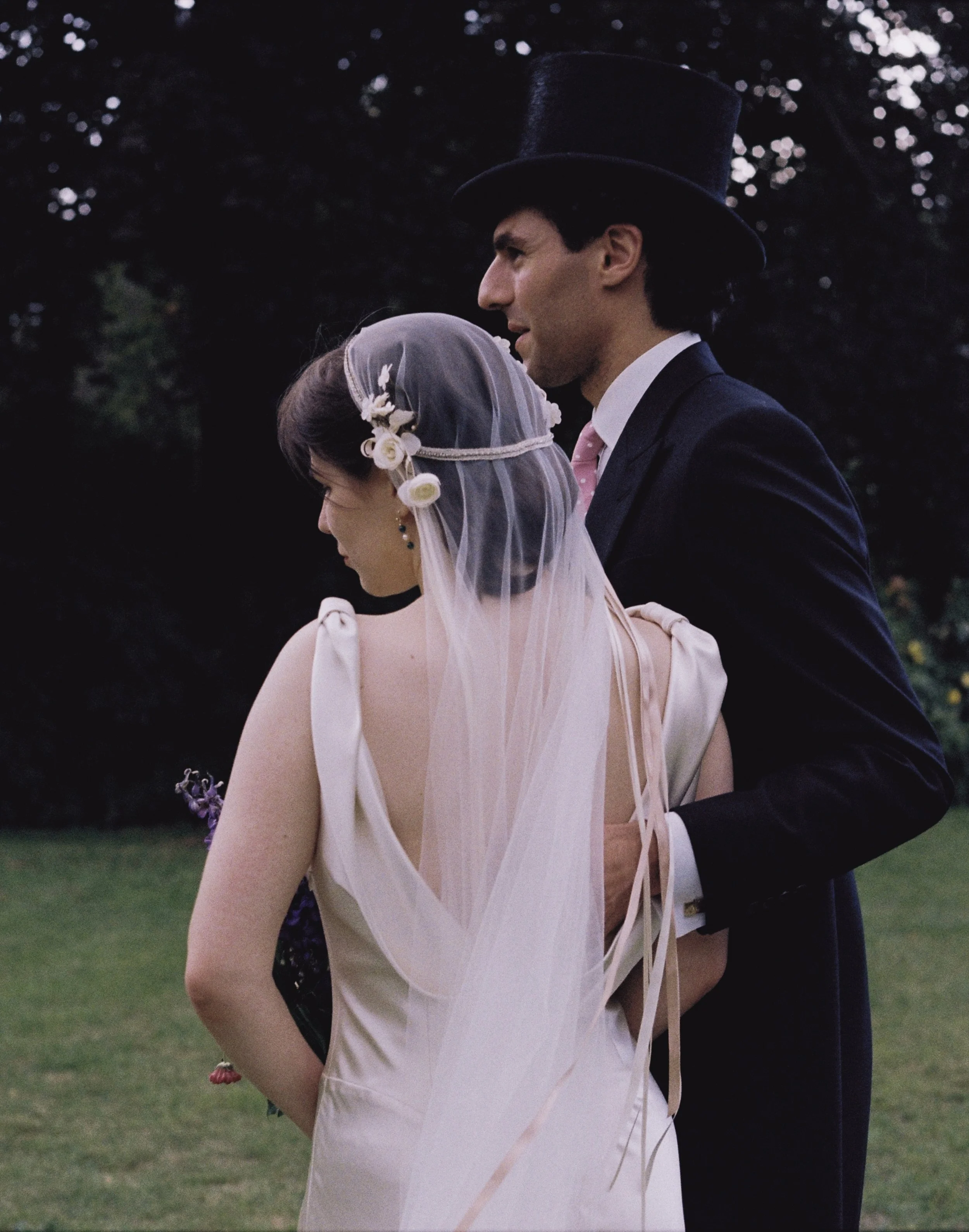 A man and woman dressed in vintage wedding attire stand outdoors during dusk, with trees in the background. The man wears a dark suit, white shirt, pink tie, and a black top hat, while the woman wears a white wedding gown with a veil, and has floral 