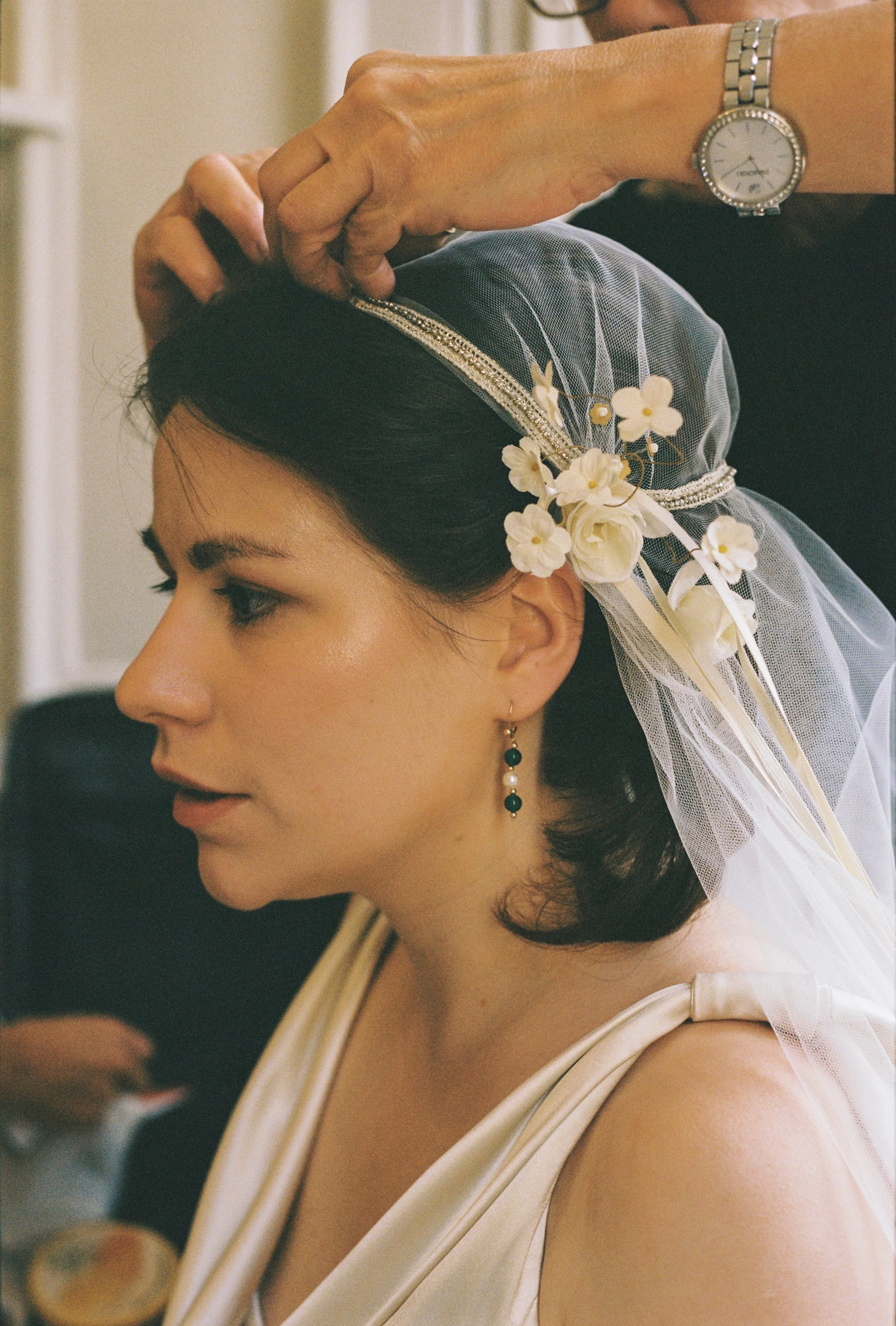 Woman with dark hair getting ready for her wedding, wearing a Juliet Cap wedding veil with white floral decorations, a satin dress, and black and gold earrings. An older person with a watch is adjusting her veil.
