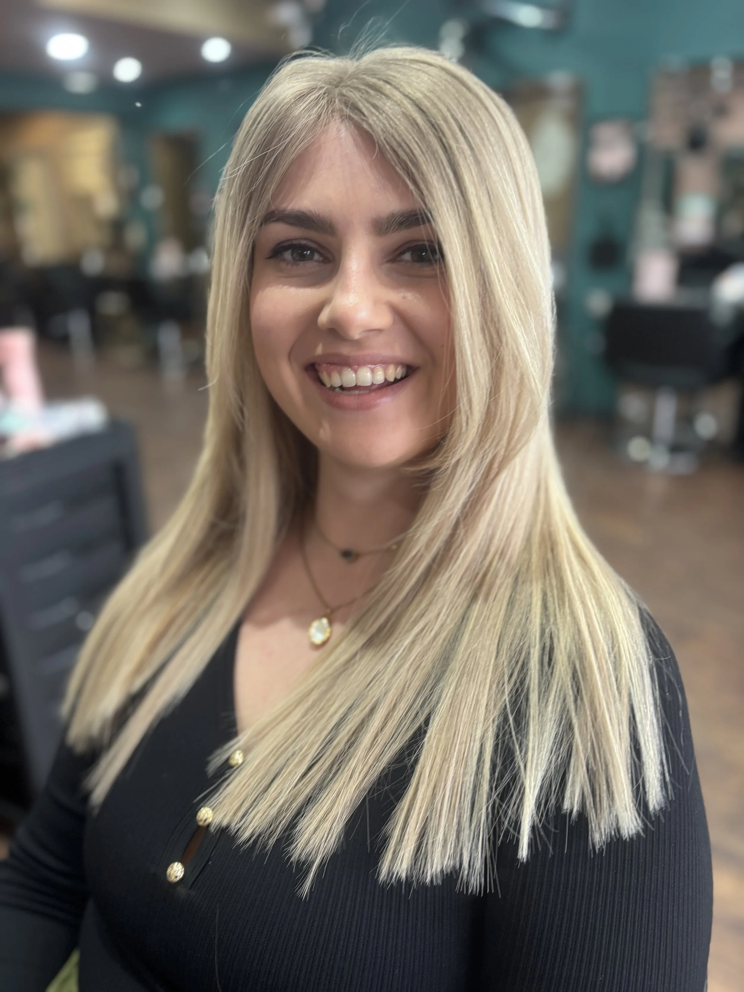 A smiling woman with long blonde hair wearing a black top and layered necklaces, standing in a hair salon.