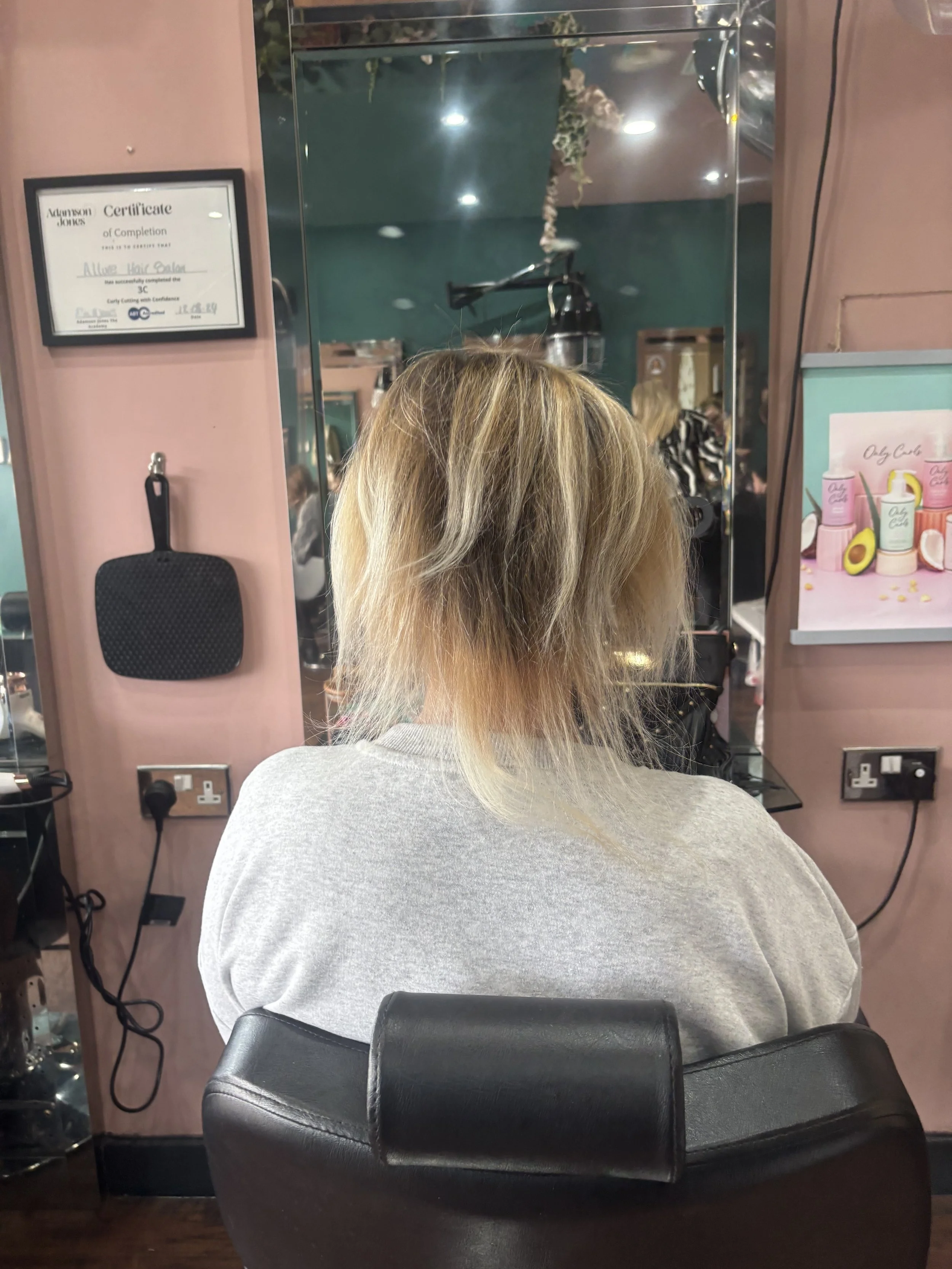 Back view of a woman with blonde, slightly messy hair sitting in a salon chair, facing a mirror, with a pink wall and salon equipment visible in the background.