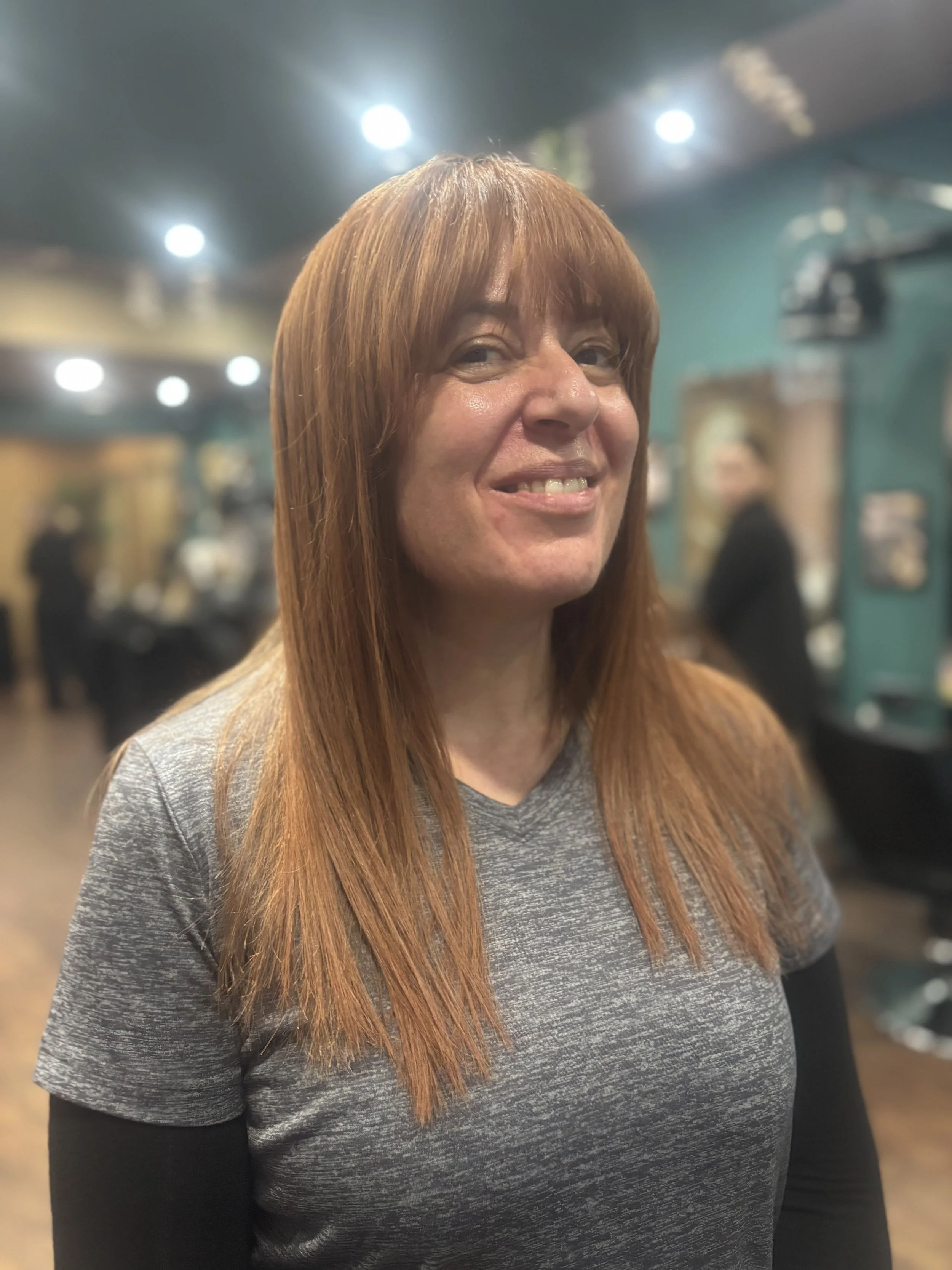 A woman with long red hair smiling in a salon or barbershop, with people and salon chairs in the background.