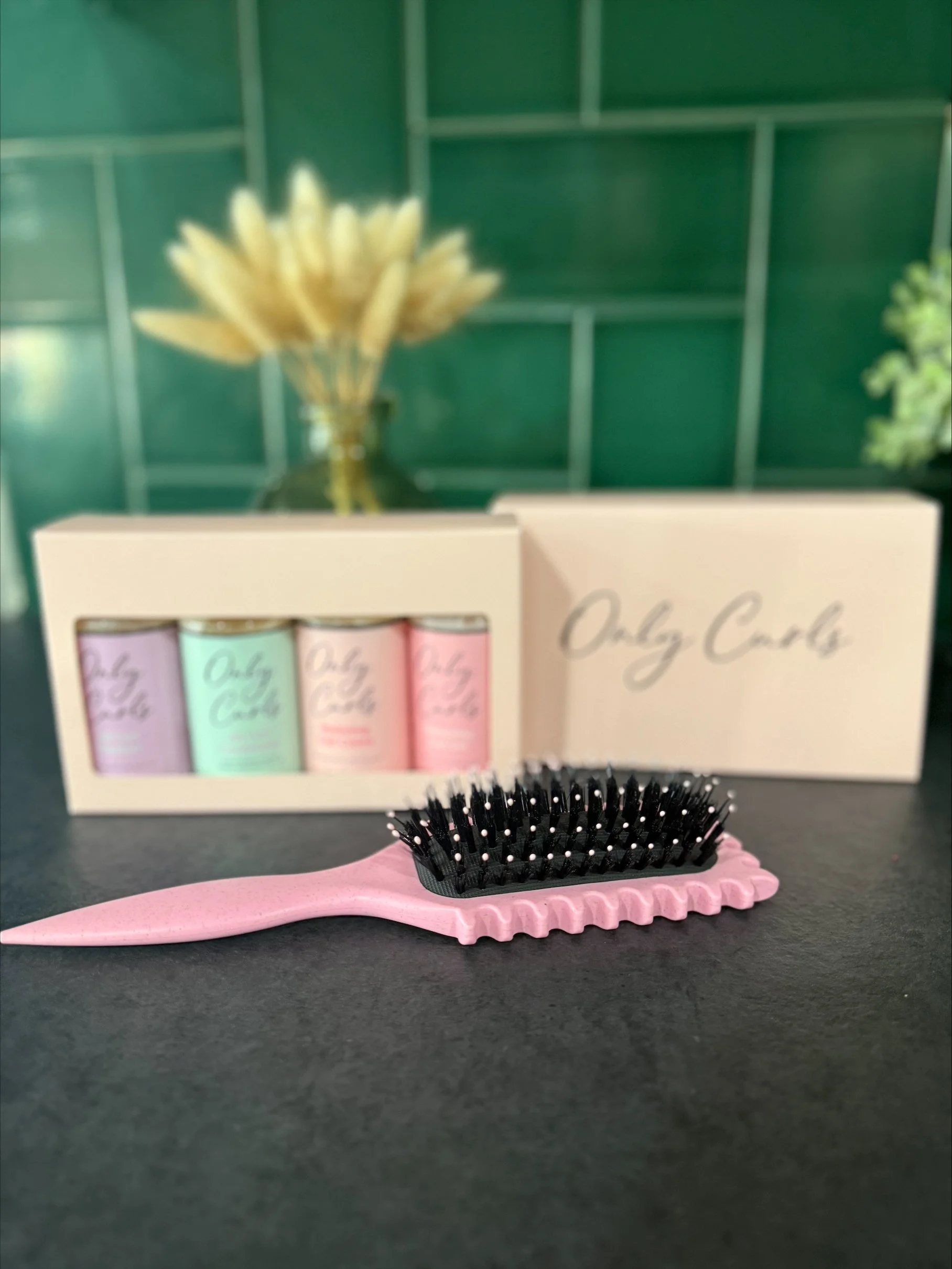 A pink hairbrush with black bristles rests on a black surface in front of a box and a set of pastel-colored nail care products. The background features a blurred green wall and a vase with dried flowers.