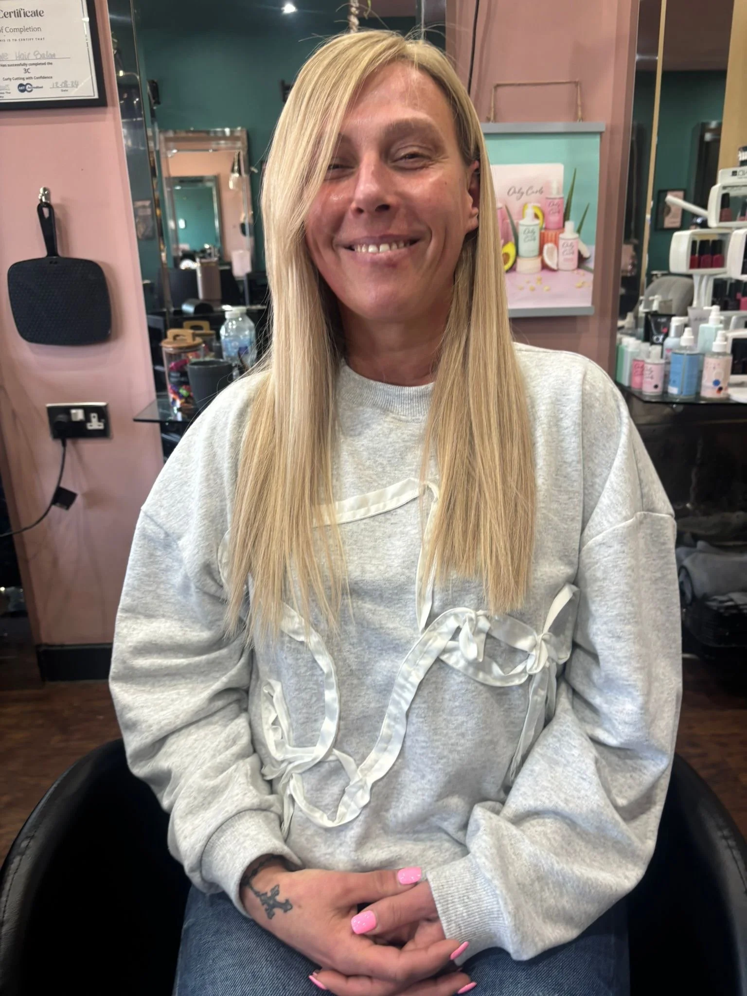 A smiling woman with long blonde hair sitting in a salon chair, wearing a gray sweatshirt with white trim and a bow detail, inside a hair salon with various hair and beauty products in the background.