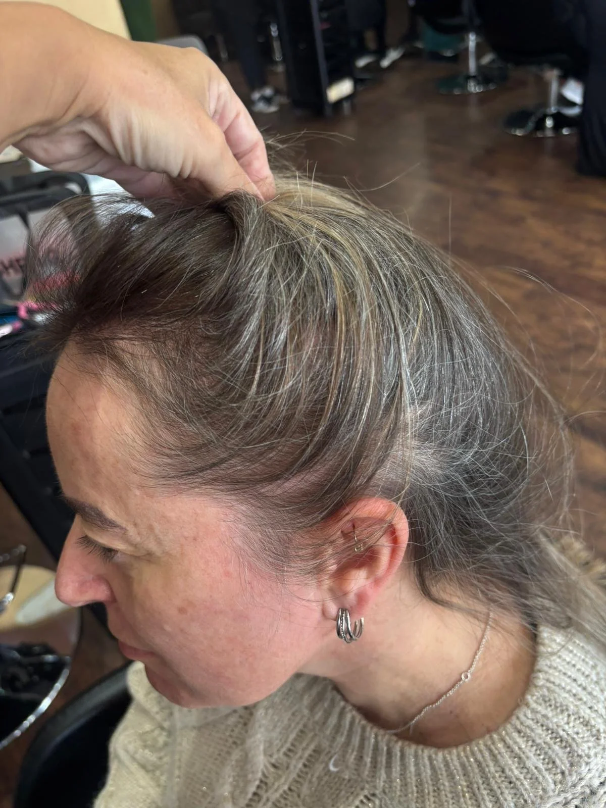 Close-up of a woman at a hair salon with gray and blonde hair, getting her hair styled. A person is lifting her hair with their hand. The woman is wearing earrings, a necklace, and a beige sweater. The salon has wooden flooring and black chairs.