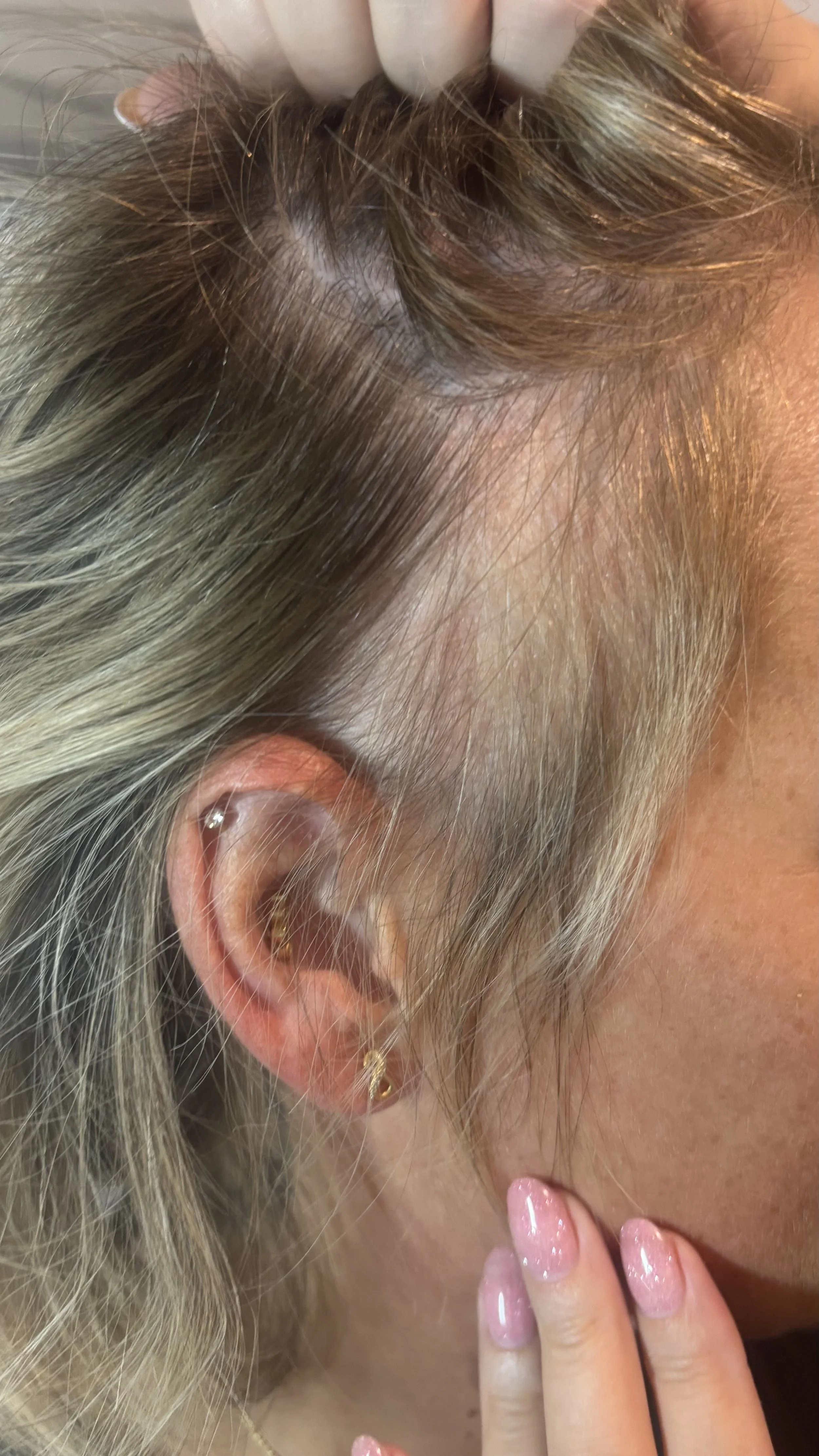 Close-up of a woman's ear with multiple earrings and a hand with painted nails near her face. She has blonde hair and light skin.