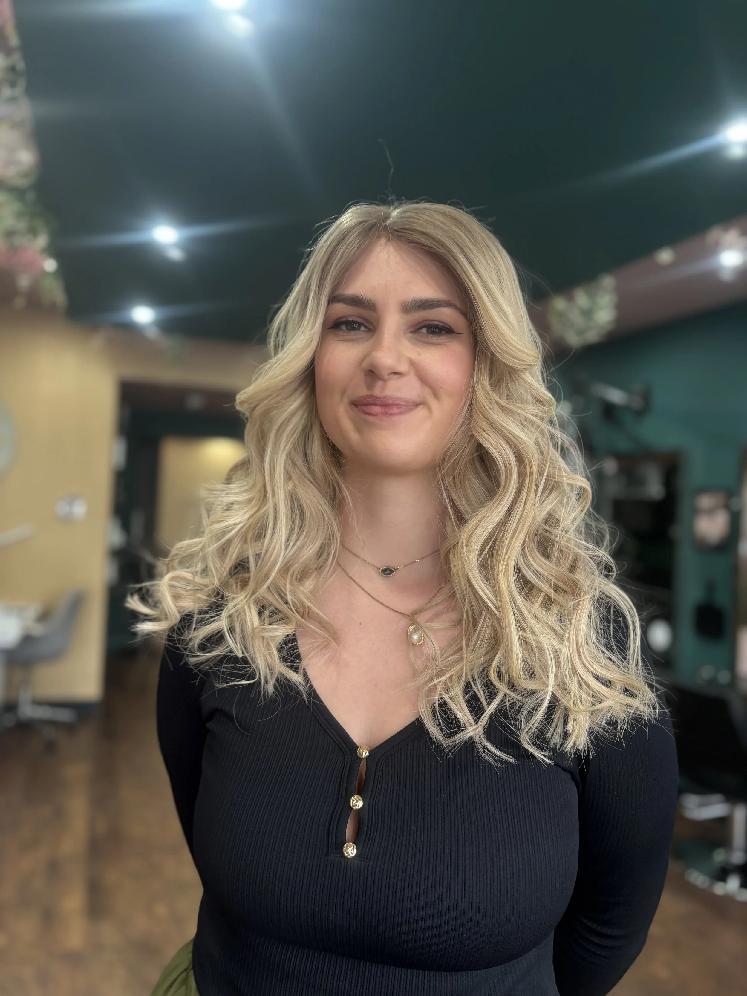 A young woman with long blonde hair styled in loose waves, smiling and standing indoors with a blurred background featuring a green ceiling, lighting, and some furniture.