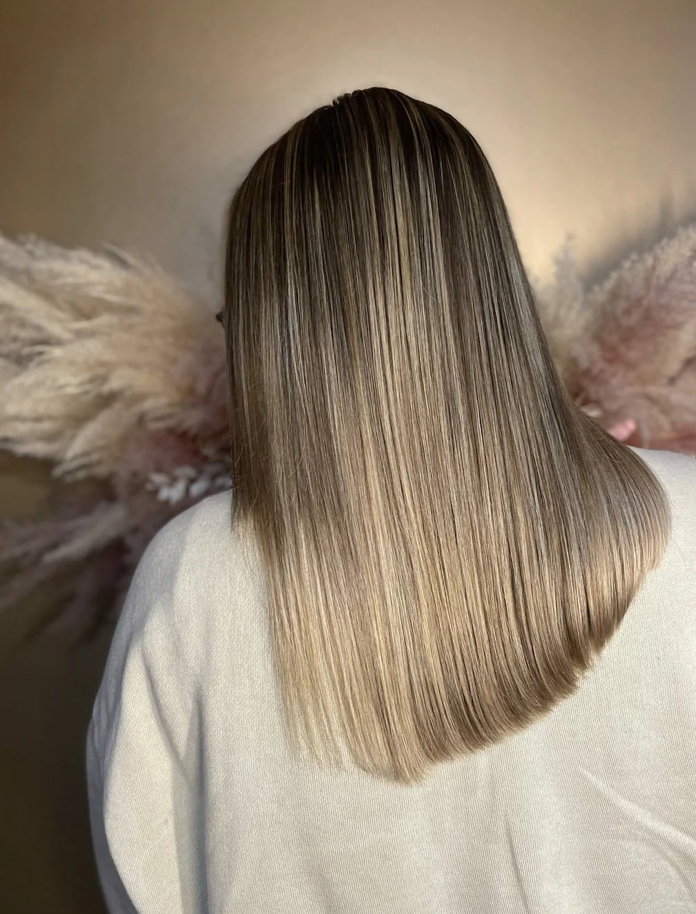 Back of a person with straight, shiny, blonde hair, wearing a white top, with a blurred pink and beige decorative background.