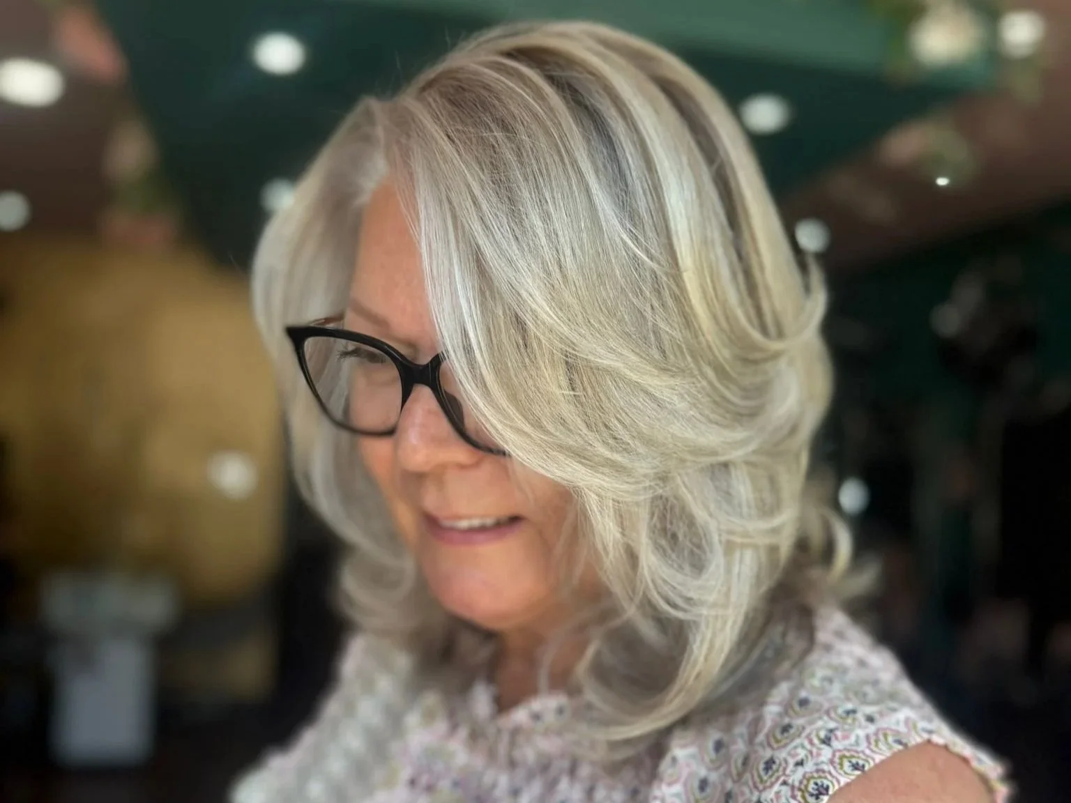 A woman with blonde, curly, shoulder-length hair wearing black eyeglasses, smiling, in an indoor setting.