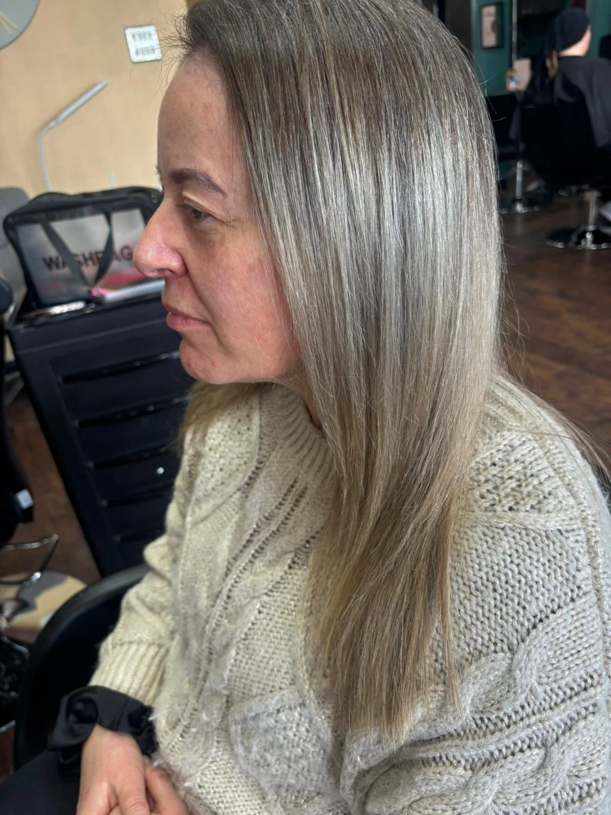 Woman with long, straight, blonde hair sitting in a salon chair, wearing a beige knit sweater, in a hair salon environment.