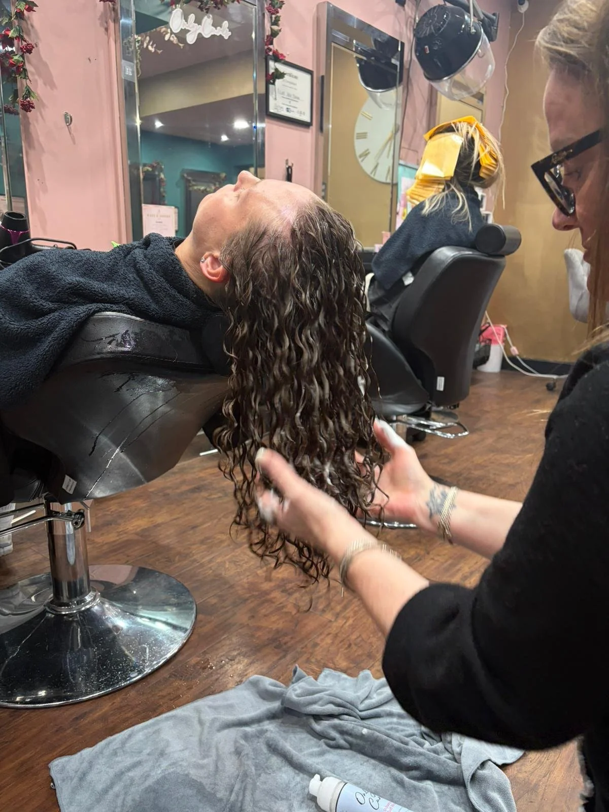 A woman is lying back in a salon chair with her head tilted, while a stylist gently washes her long, curly hair. The salon has pink walls, mirrors, and various hairdressing tools visible in the background.