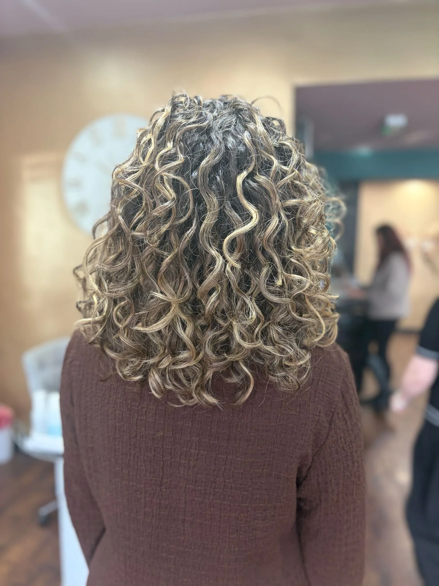 Back view of a woman with shoulder-length, curly, blonde-highlighted hair in a hair salon.