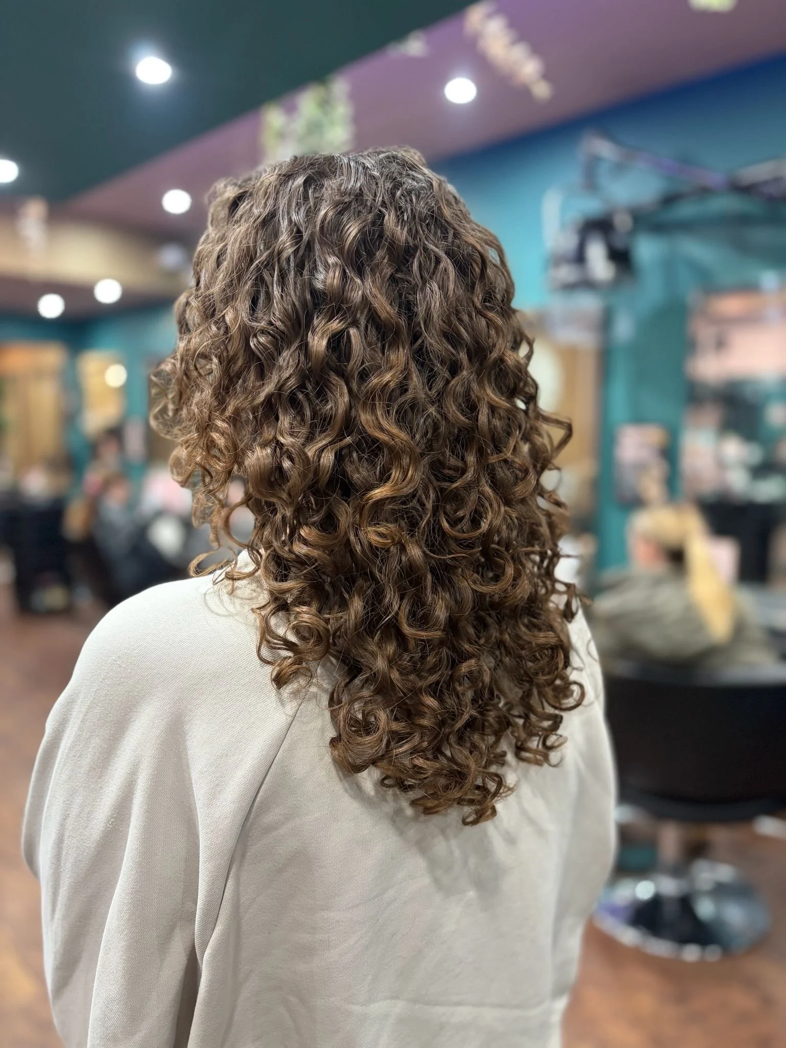 Back view of a person with long, curly brown hair in a salon or similar setting.