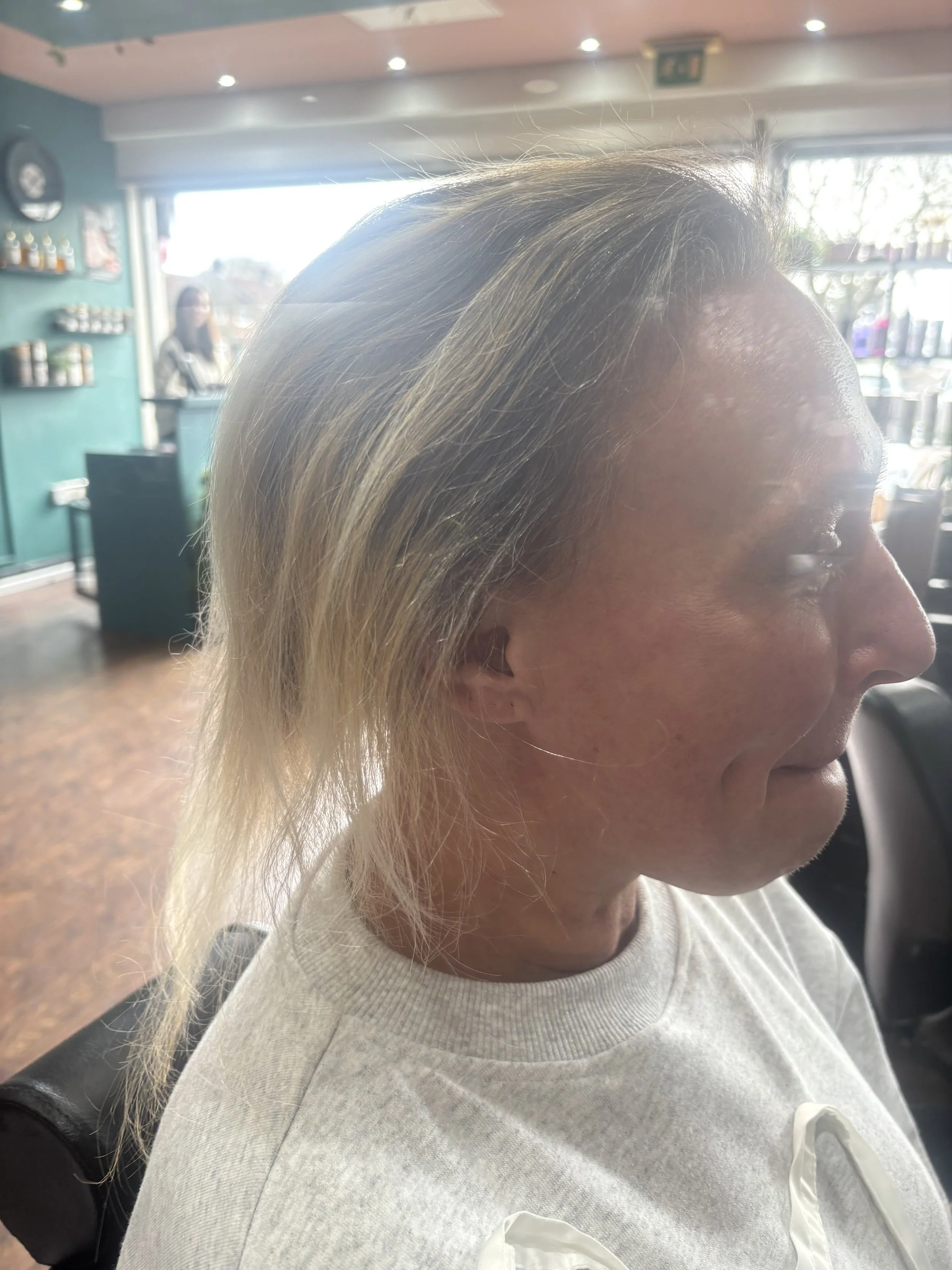 Woman with blonde hair in a salon, sitting in a chair, with her profile visible against a bright window and a teal wall in the background.