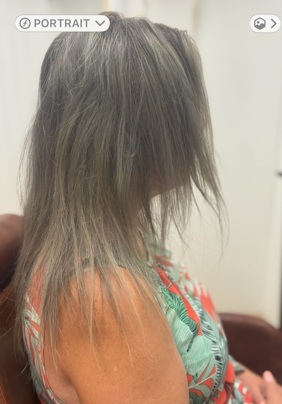 Side view of a woman with tousled, shoulder-length light brown or blonde hair, wearing a colorful, patterned sleeveless top, sitting on a brown chair in a room with cream-colored walls.