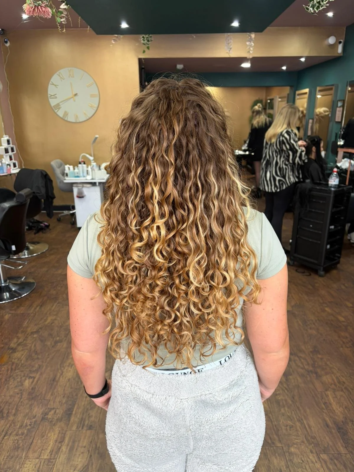 Back view of a woman with long, blonde, curly hair in a salon with other stylists and clients, a large wall clock, and salon equipment.