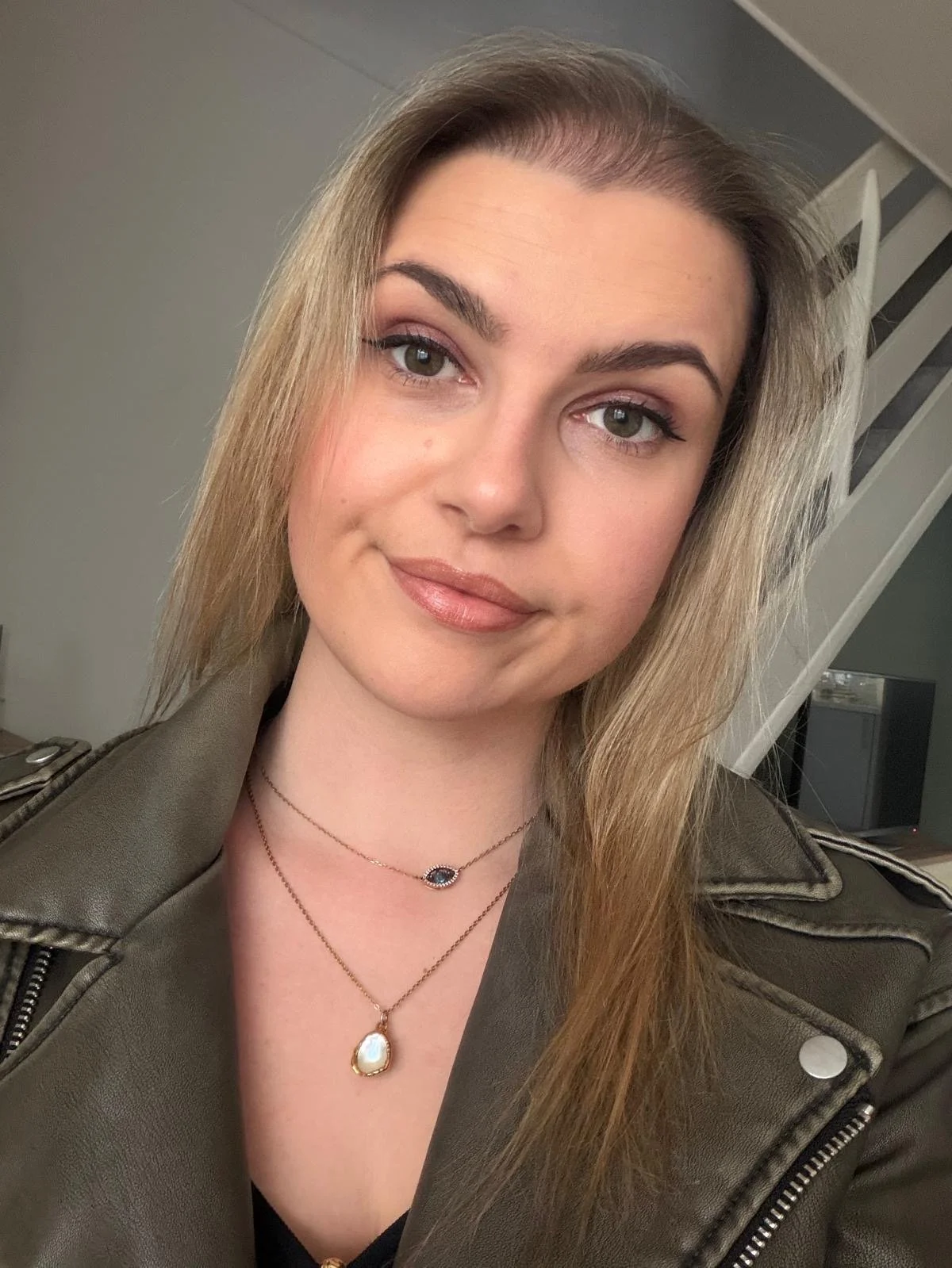 Close-up portrait of a young woman with light brown hair, wearing a olive green jacket, layered necklaces, and makeup, smiling indoors.