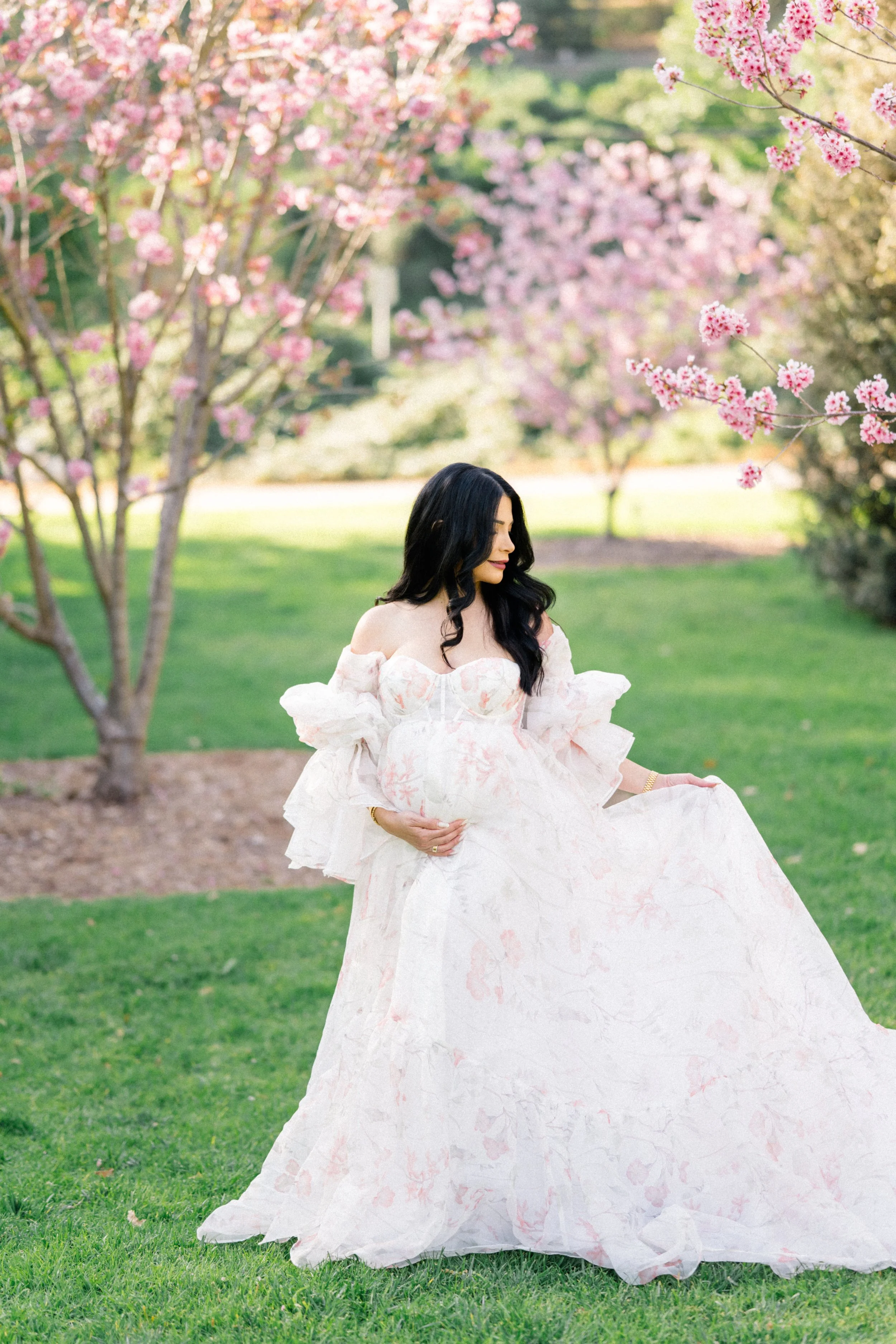 Cherry blossom Maternity photos of an expecting mother standing beneath blooming pink trees, holding her baby bump and lifting her flowing floral gown in soft, natural spring light.
