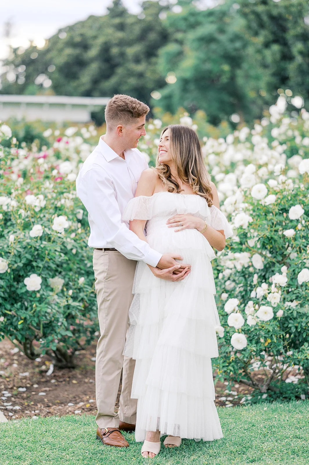 Summer Maternity Photos of an expecting couple embracing in a blooming rose garden, with the mother wearing a soft white dress surrounded by white flowers and greenery.