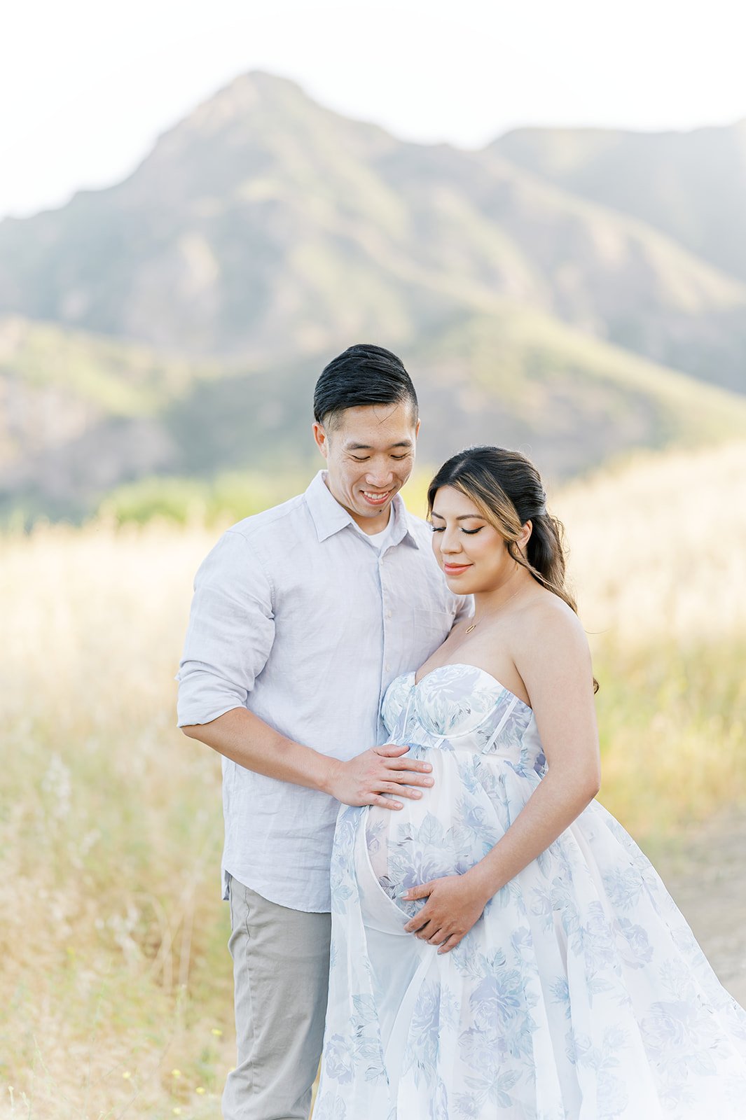 Summer Maternity Photos of an expecting couple standing in a golden mountain field, with the mother wearing a soft blue floral gown during golden hour.