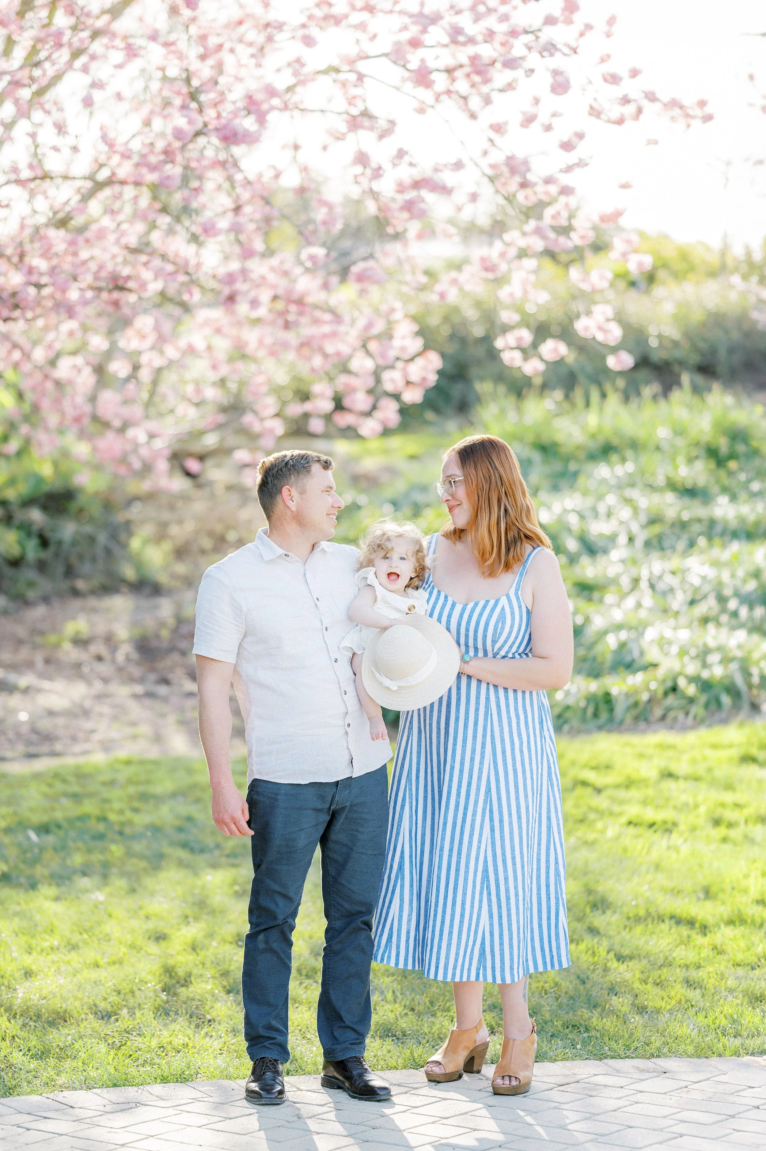 Cherry Blossom Photoshoot ideas featuring a family standing together beneath blooming cherry blossom trees, with parents holding their smiling child and soft sunlight creating a bright, airy spring atmosphere