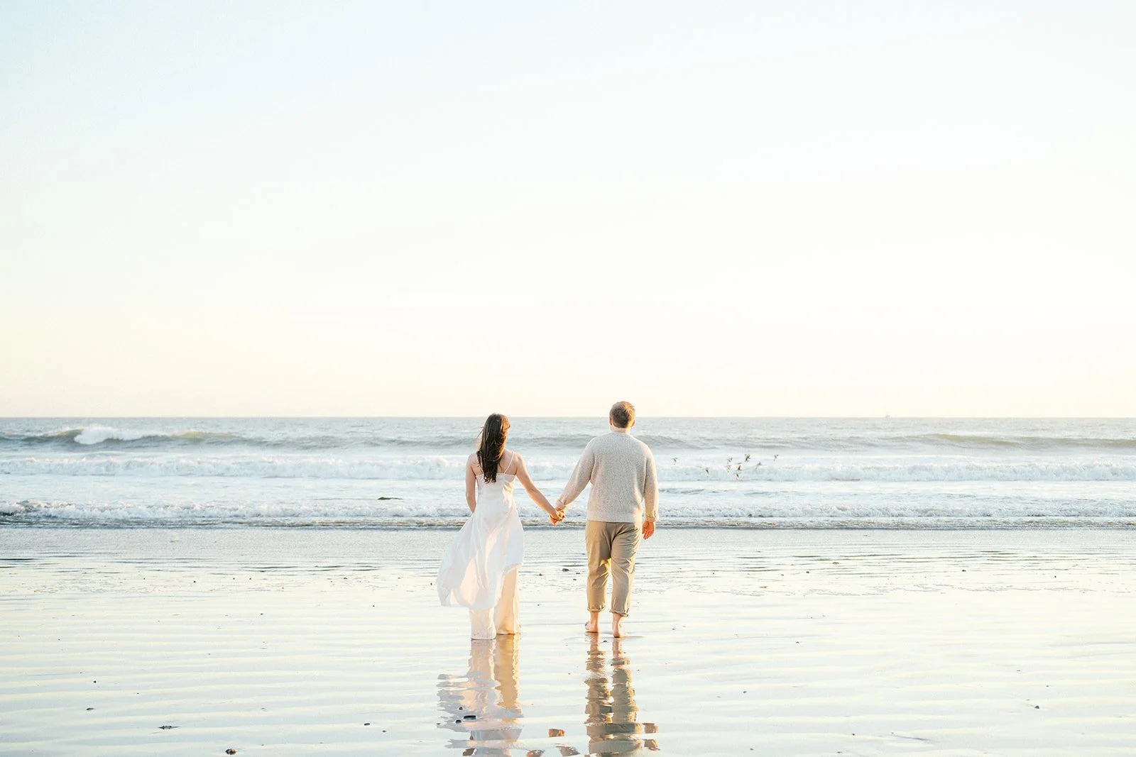 A couple walks hand-in-hand along the shoreline at sunset, their reflections visible on the wet sand as gentle waves roll in behind them on a calm Malibu beach.
