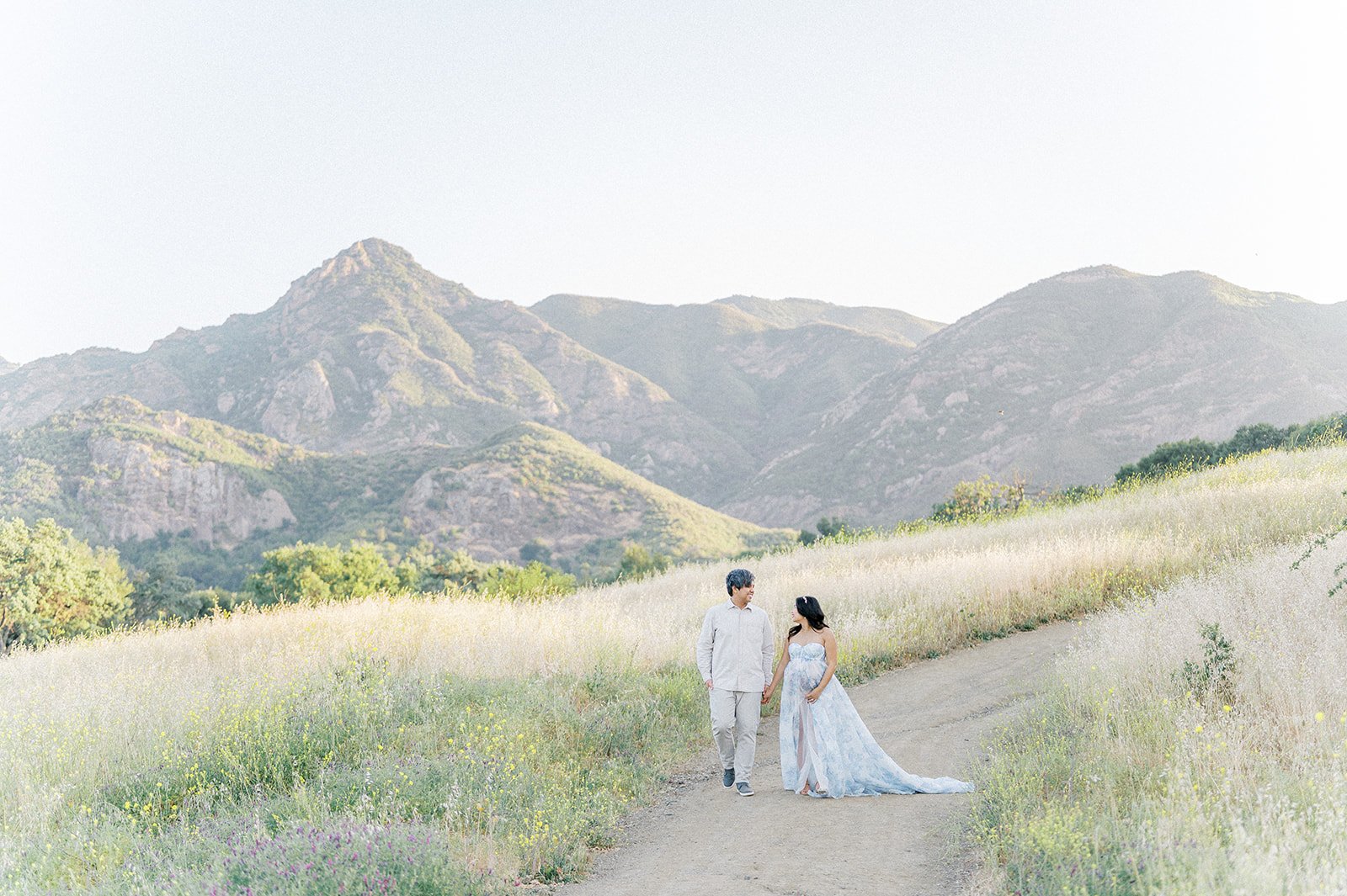 Expectant couple walks hand in hand along a dirt path surrounded by wildflowers and golden grass, with sunlit green mountains rising softly in the background.