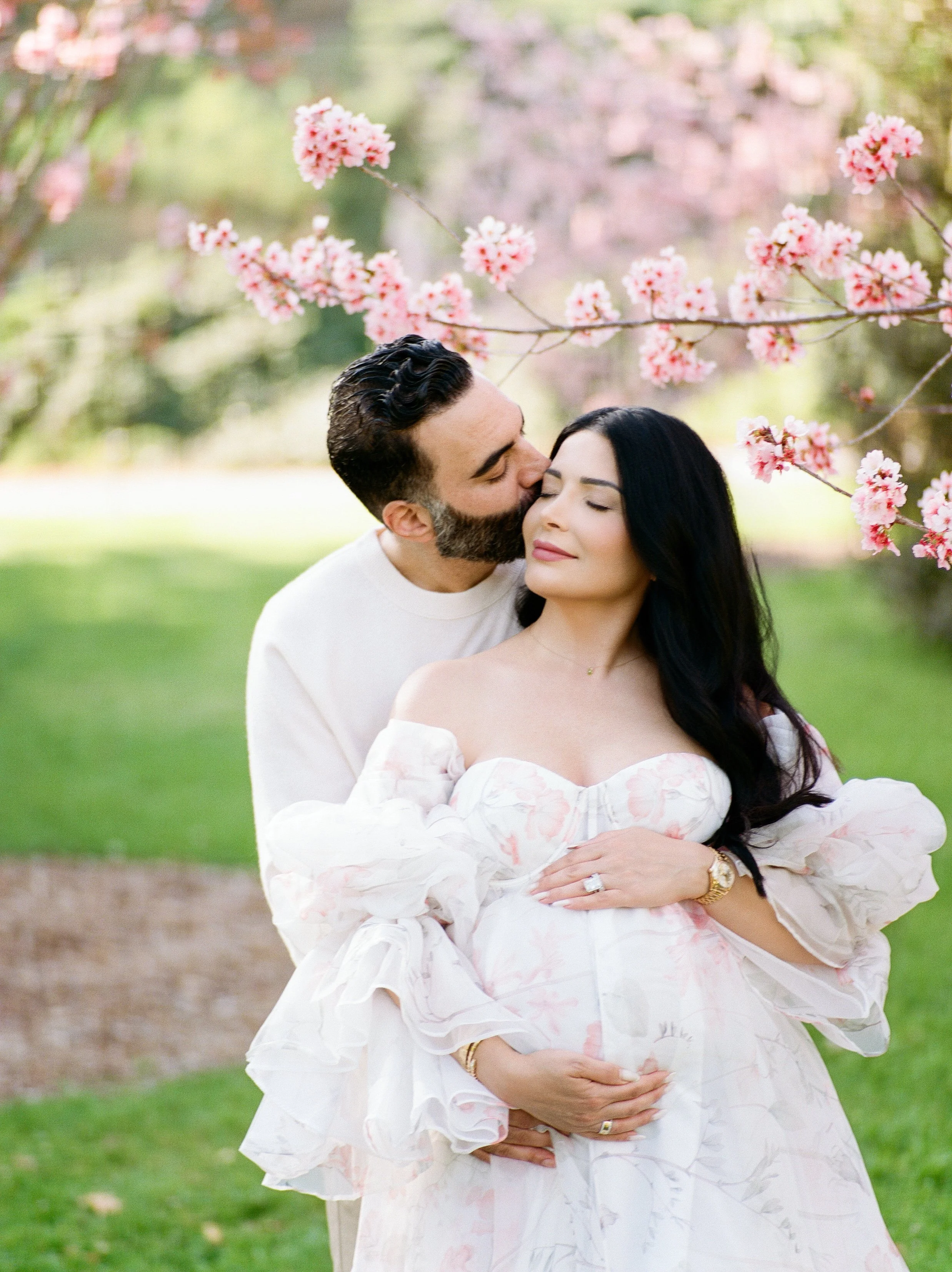 Cherry blossom Maternity photos of an expecting couple beneath blooming pink trees, with the partner gently kissing the mother’s cheek as they cradle her baby bump in soft, natural spring light.