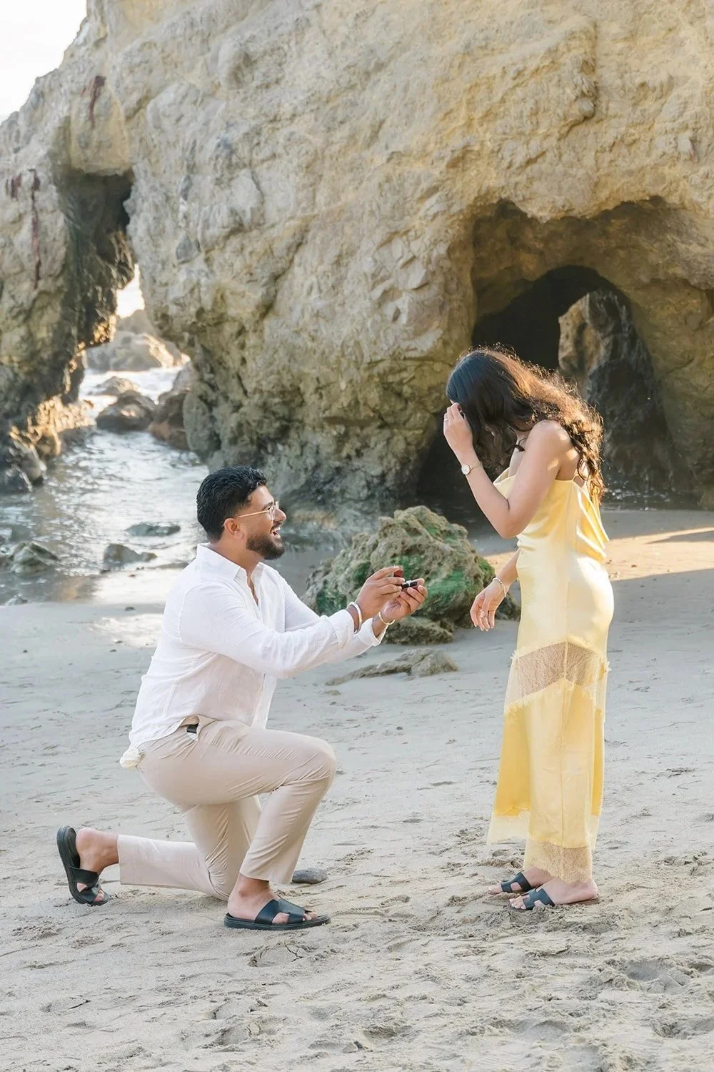 A man kneels on the sand during a New Years Eve Proposal in Malibu, presenting a ring to his partner on a quiet beach framed by coastal rock formations and soft winter light.