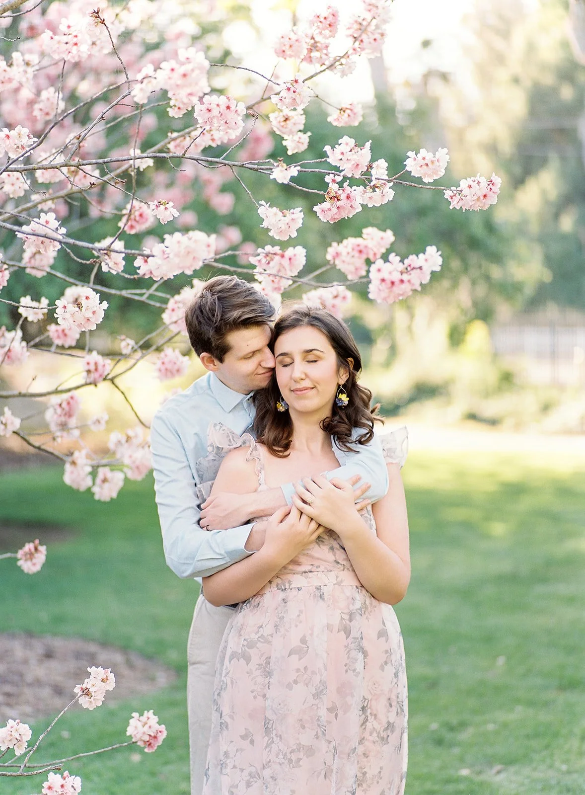 Cherry Blossom Photoshoot ideas capturing a couple in a soft embrace beneath blooming cherry blossom branches, with eyes closed and gentle expressions as warm sunlight filters through the pastel pink flowers