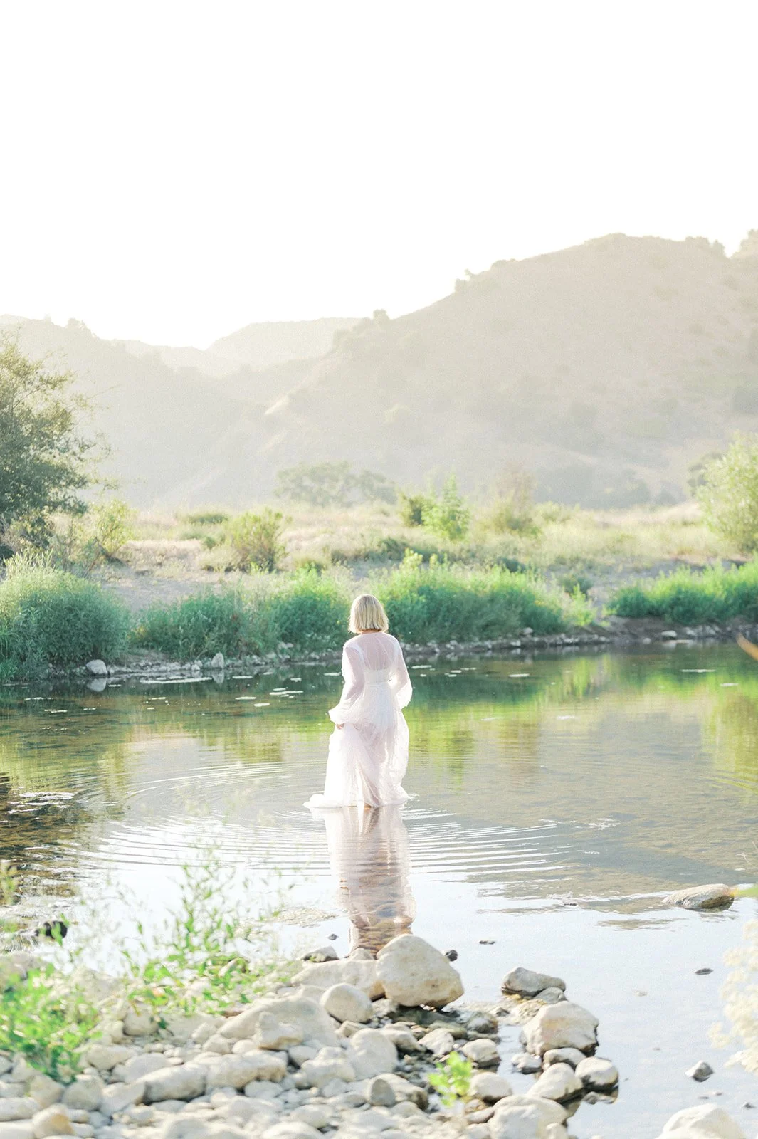 Summer Maternity Photos of an expecting mother in a flowing white gown standing in a calm river with soft mountain light reflecting across the water.