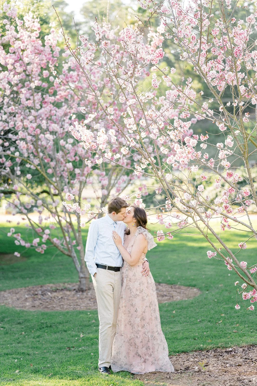 Cherry Blossom Photoshoot ideas featuring a couple sharing a kiss beneath blooming cherry blossom trees, surrounded by soft pink flowers in a light, airy spring setting