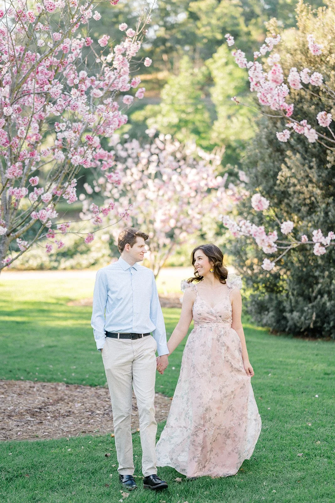 Cherry Blossom Photoshoot ideas showing a couple walking hand in hand beneath blooming cherry blossom trees, smiling at each other as soft pink flowers frame the scene in warm, natural light