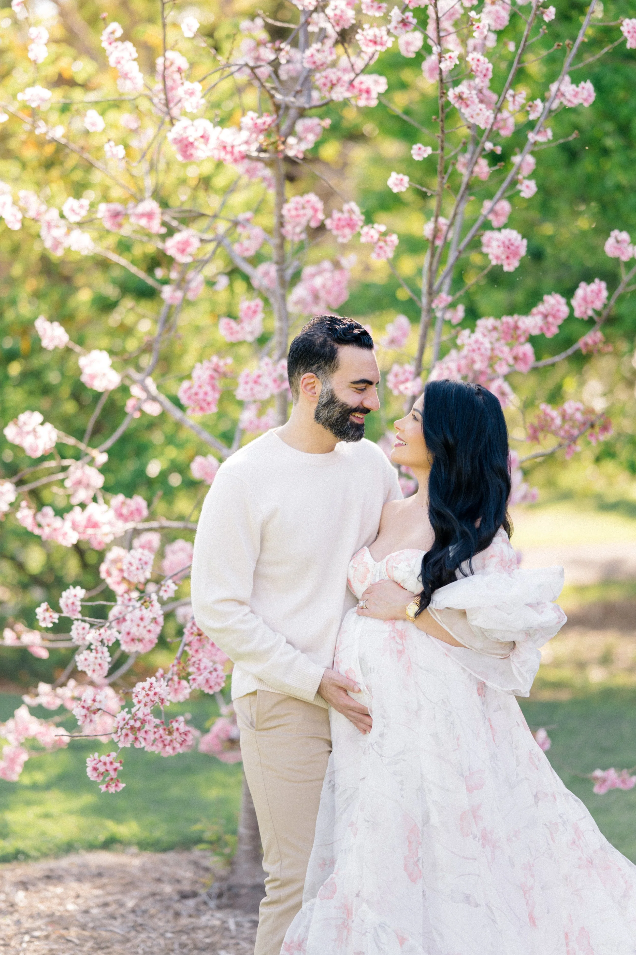 Cherry blossom Maternity photos of an expecting couple standing beneath blooming pink trees, smiling at each other while gently holding the baby bump in soft, natural spring light.