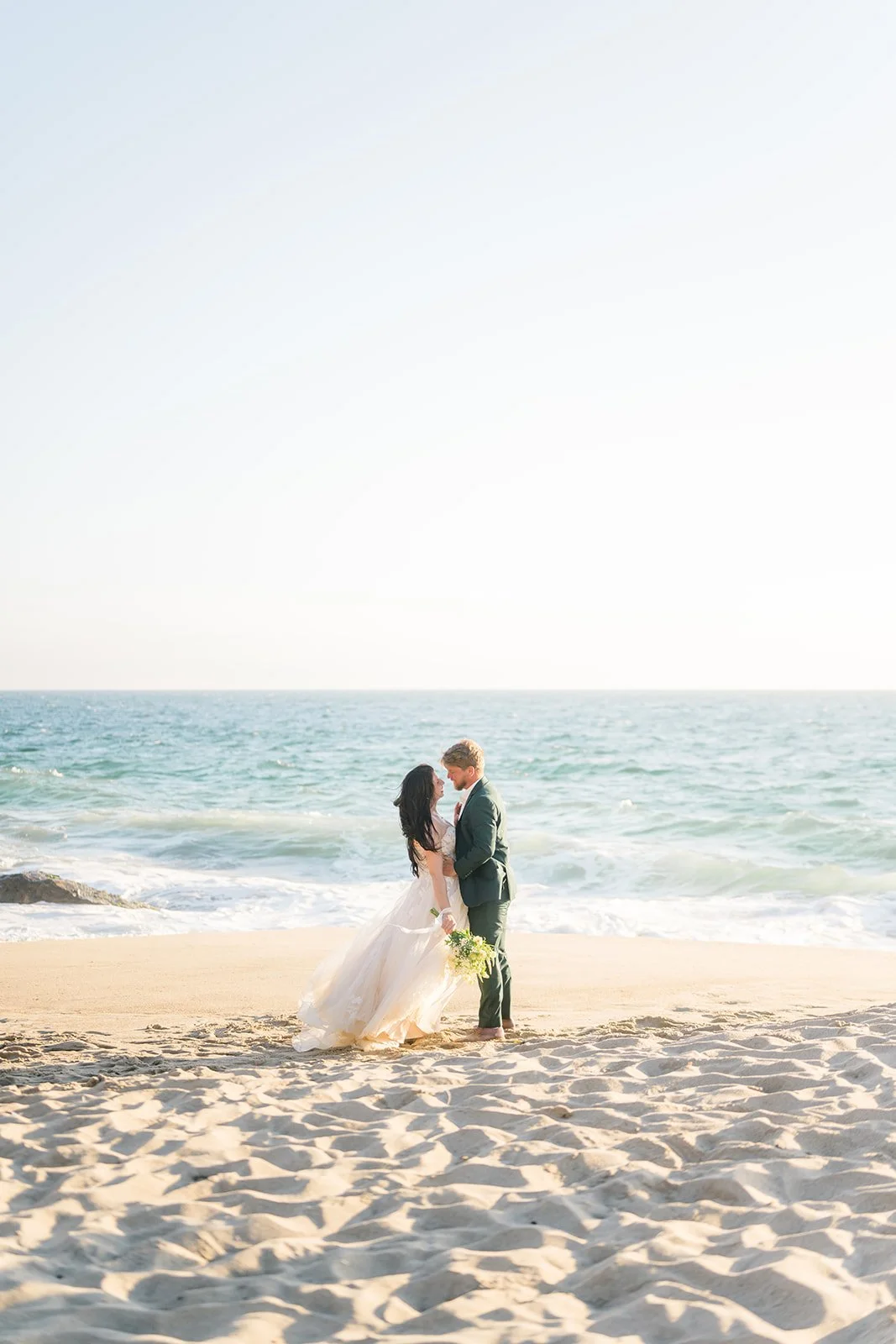 Bride and groom share a kiss on the sandy shore at Point Dume, waves gently crashing behind them under soft golden light, with the bride’s flowing dress and bouquet catching the breeze.