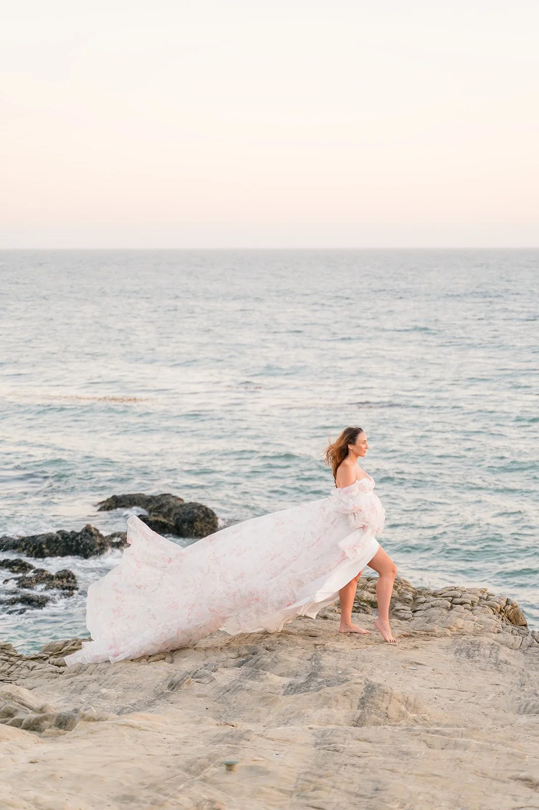 Pregnant woman in a flowing off-the-shoulder gown stands barefoot on coastal rocks at sunset, her dress billowing in the ocean breeze with soft pastel light over the sea.