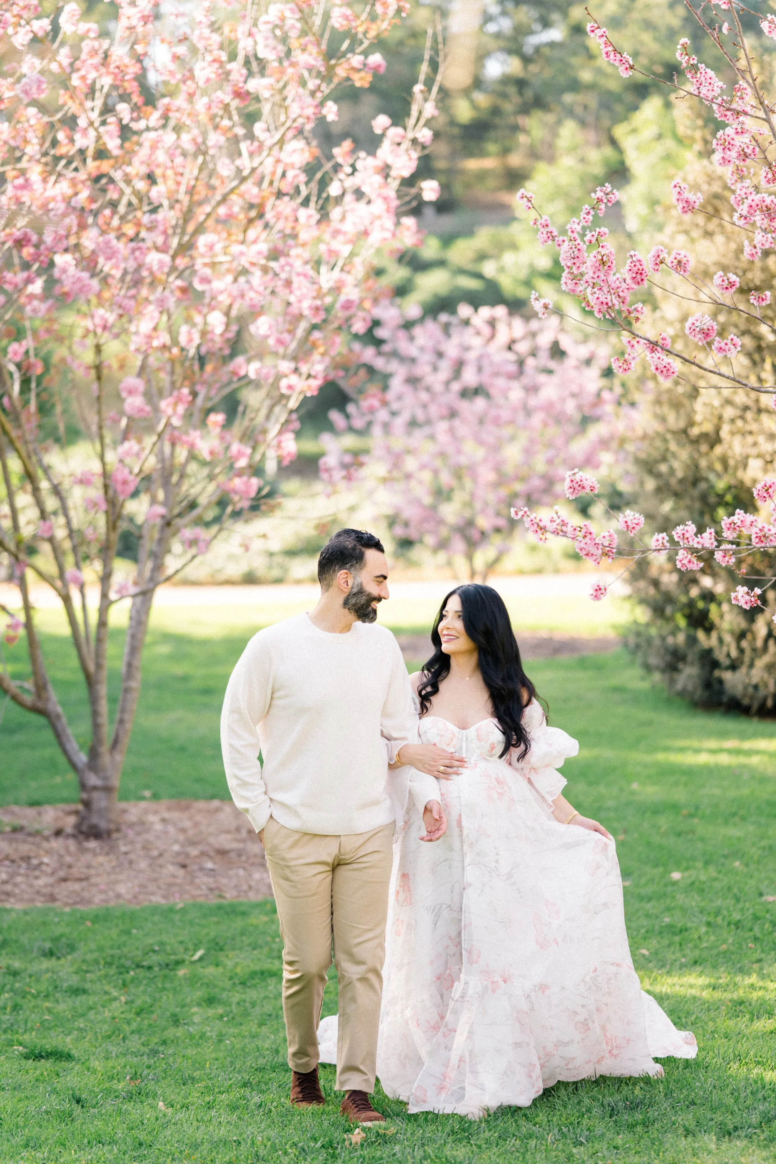 Cherry blossom Maternity photos of an expecting couple walking together beneath blooming pink trees, smiling at each other while gently holding the baby bump in soft, natural spring light.